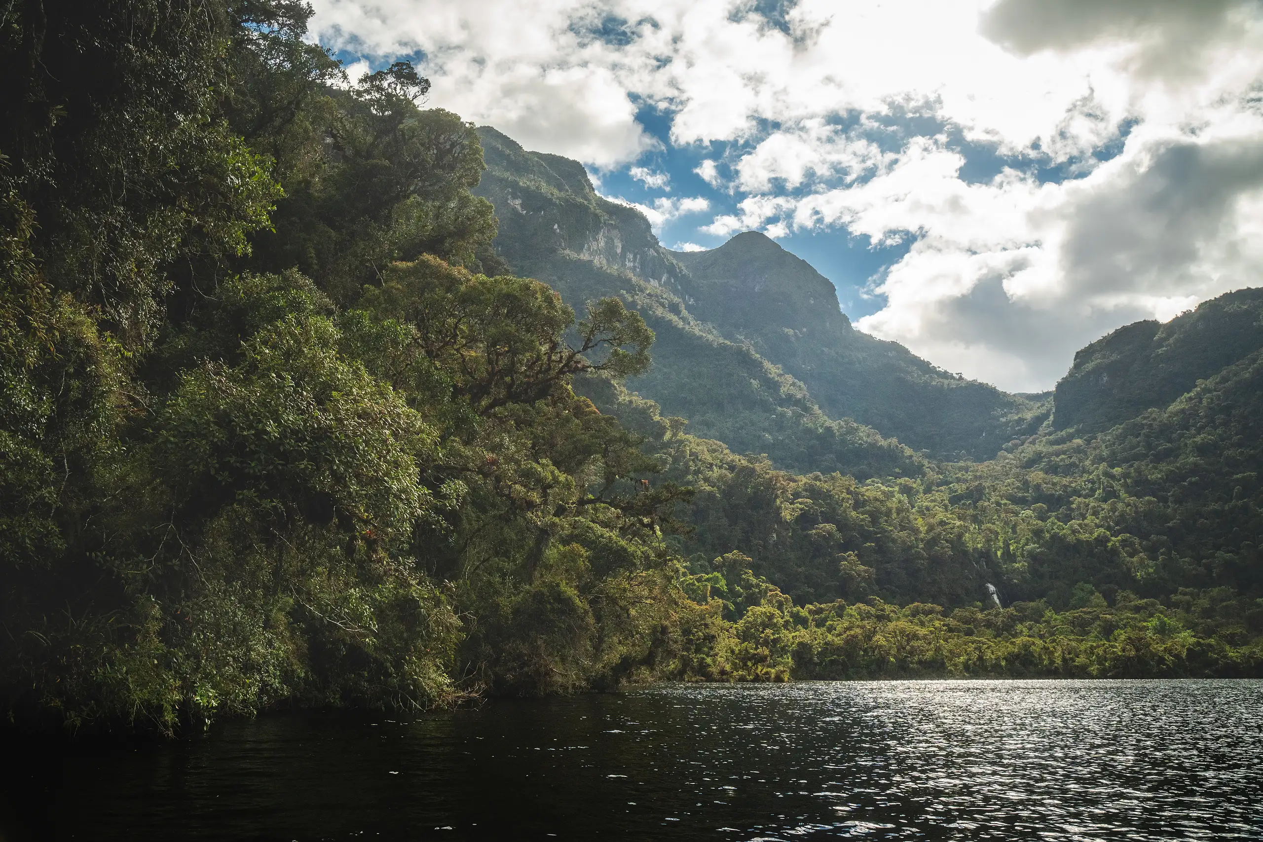 Sunlit water leading to a forested shore, steep green mountains rising on both sides of a valley under broken cloud, Laguna de los Cóndores, Amazonas, Peru