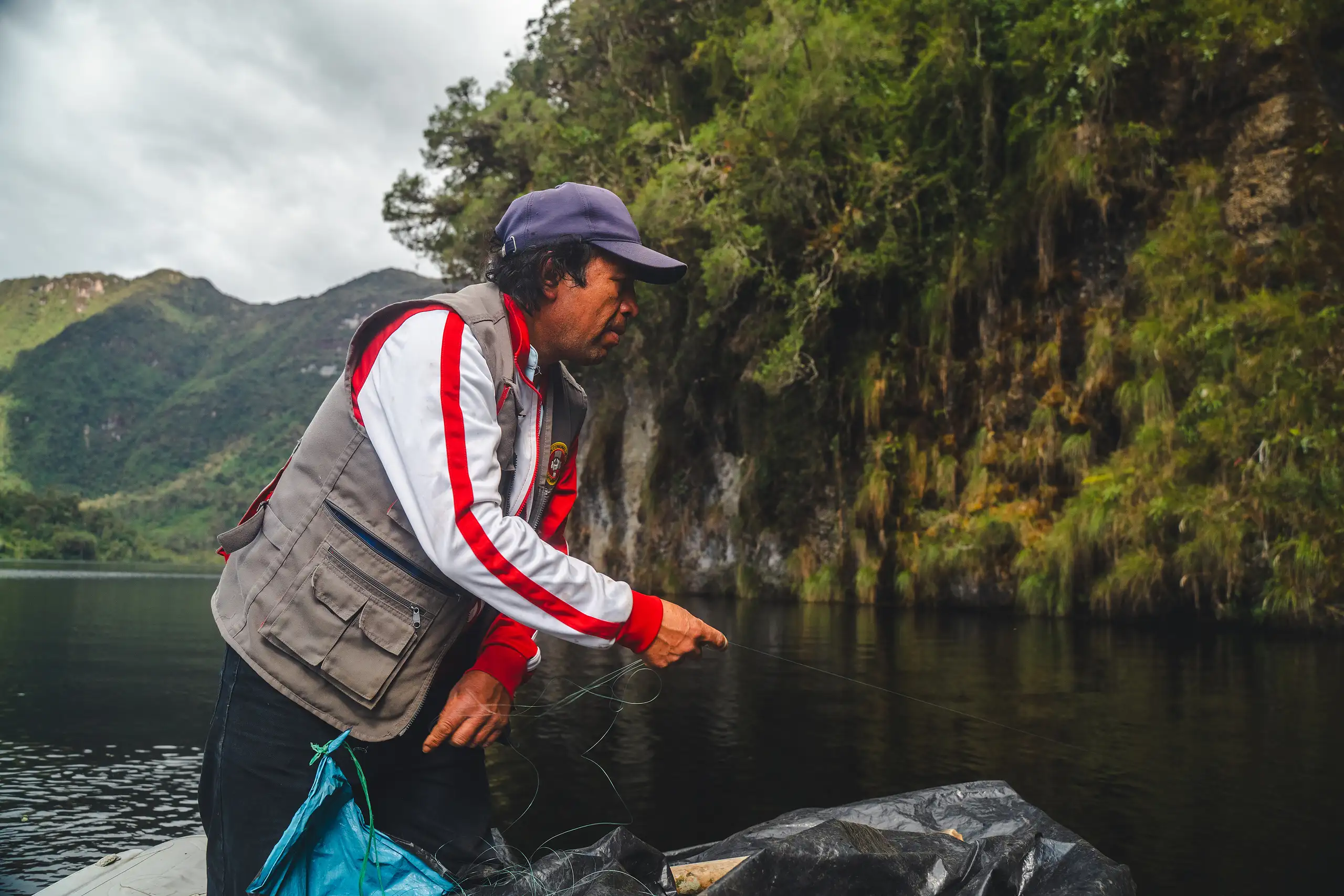 An AAV guide in a cap and a red-and-white jacket leaning over the side of a boat to handle a fishing line, a moss-covered cliff and green hills across the lake, Laguna de los Cóndores, Amazonas, Peru