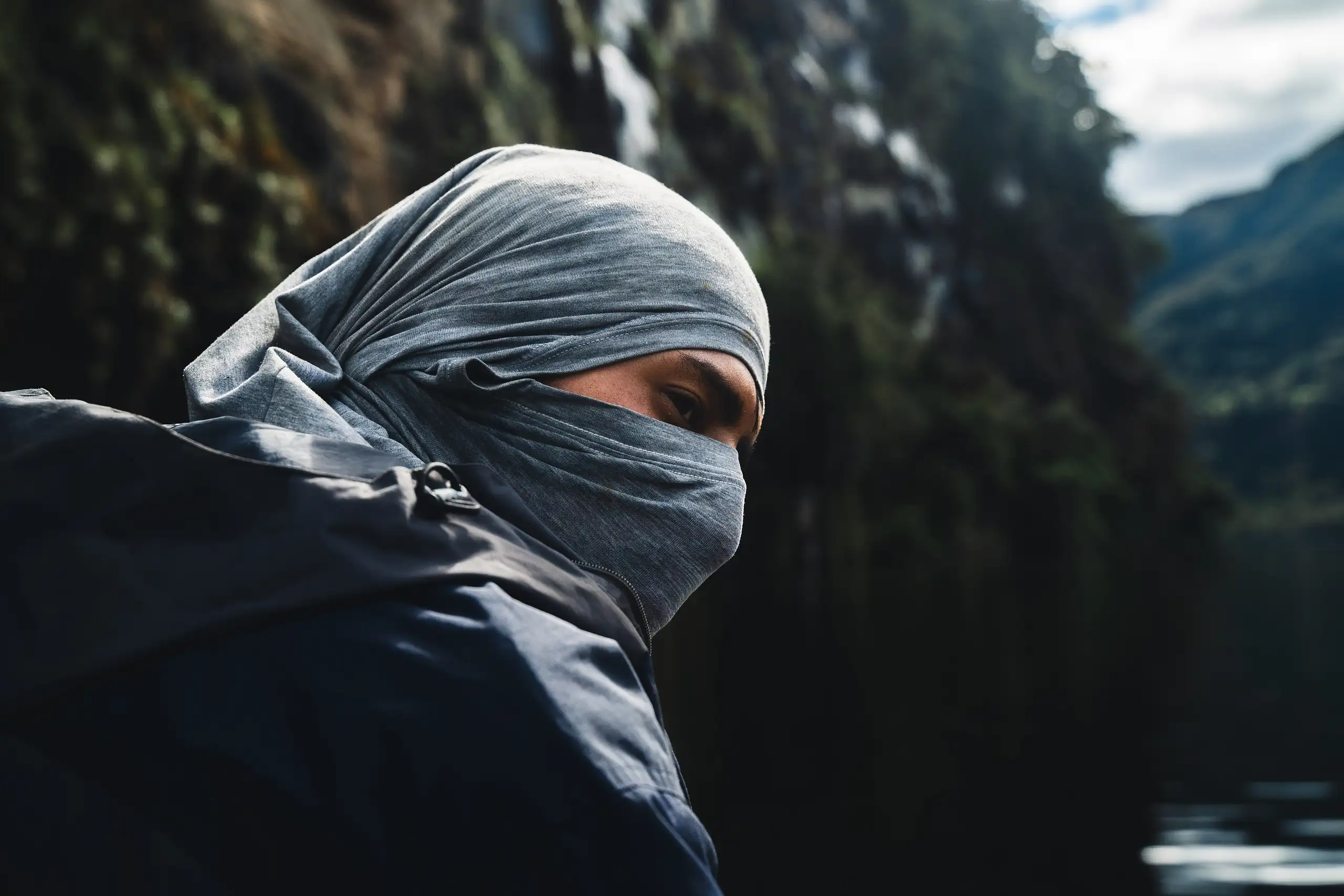 An AAV guide in a dark jacket with a grey buff pulled over their head and face, looking sideways from the boat, a dark forested cliff and water behind, Laguna de los Cóndores, Amazonas, Peru