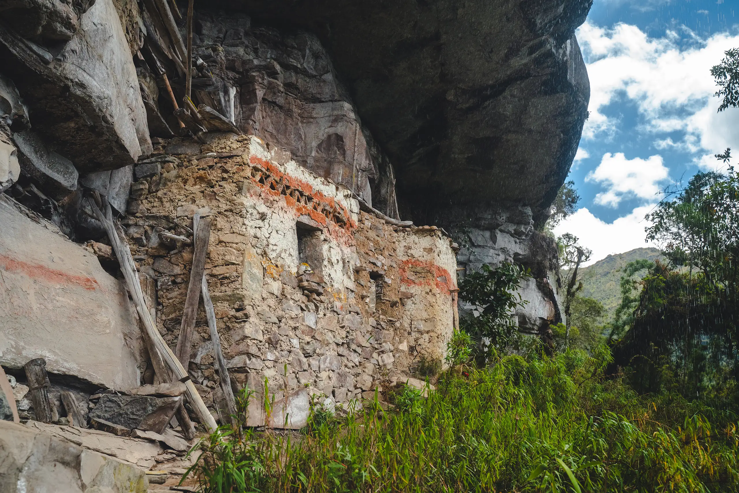 A stone funerary house with red zigzag friezes and small window openings, built under a massive rock overhang, green vegetation and blue sky beside it, Laguna de los Cóndores, Amazonas, Peru