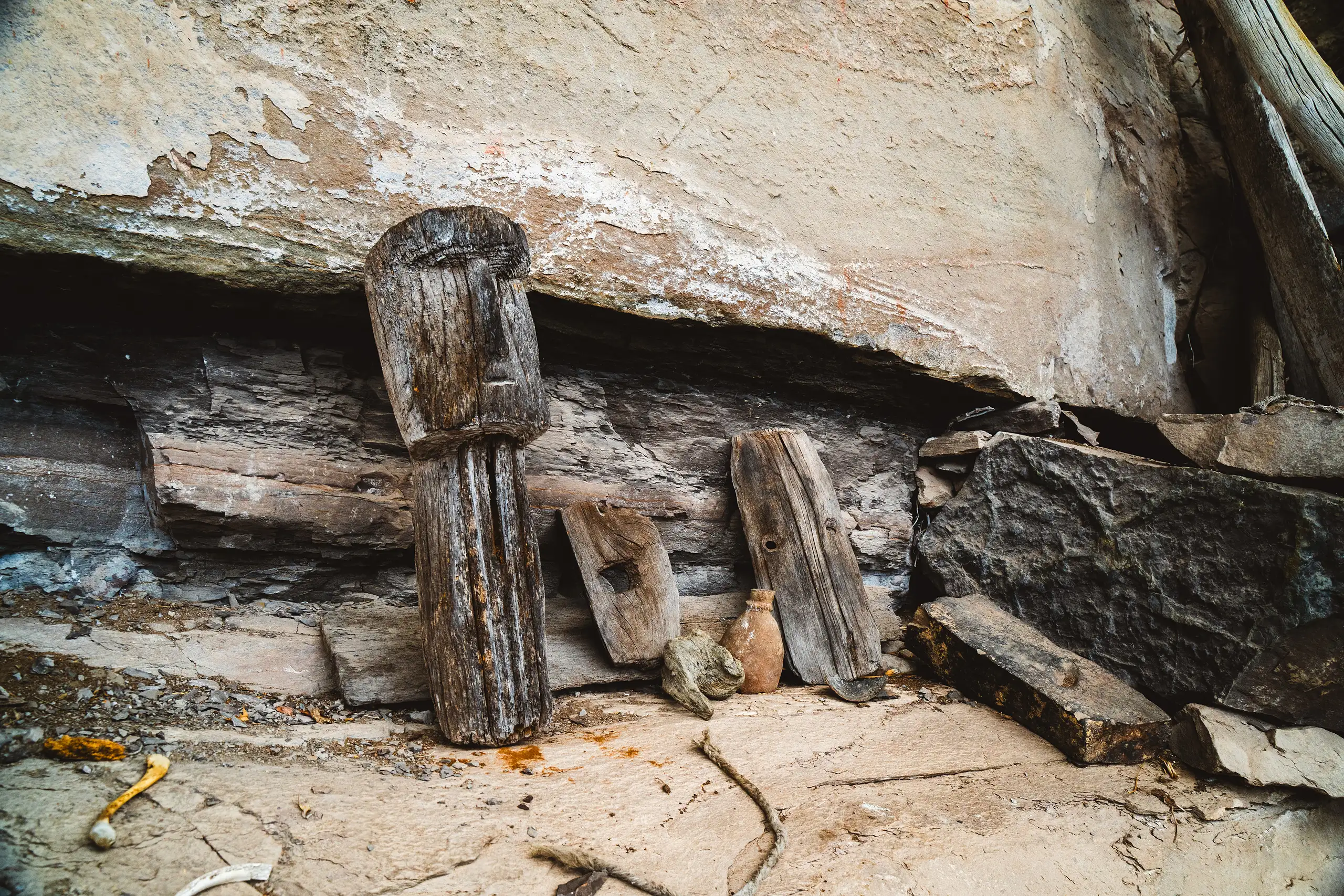 Two weathered wooden carvings with simple faces leaning against a painted rock wall beside a small clay jar, bones lying on the stone floor, Laguna de los Cóndores, Amazonas, Peru