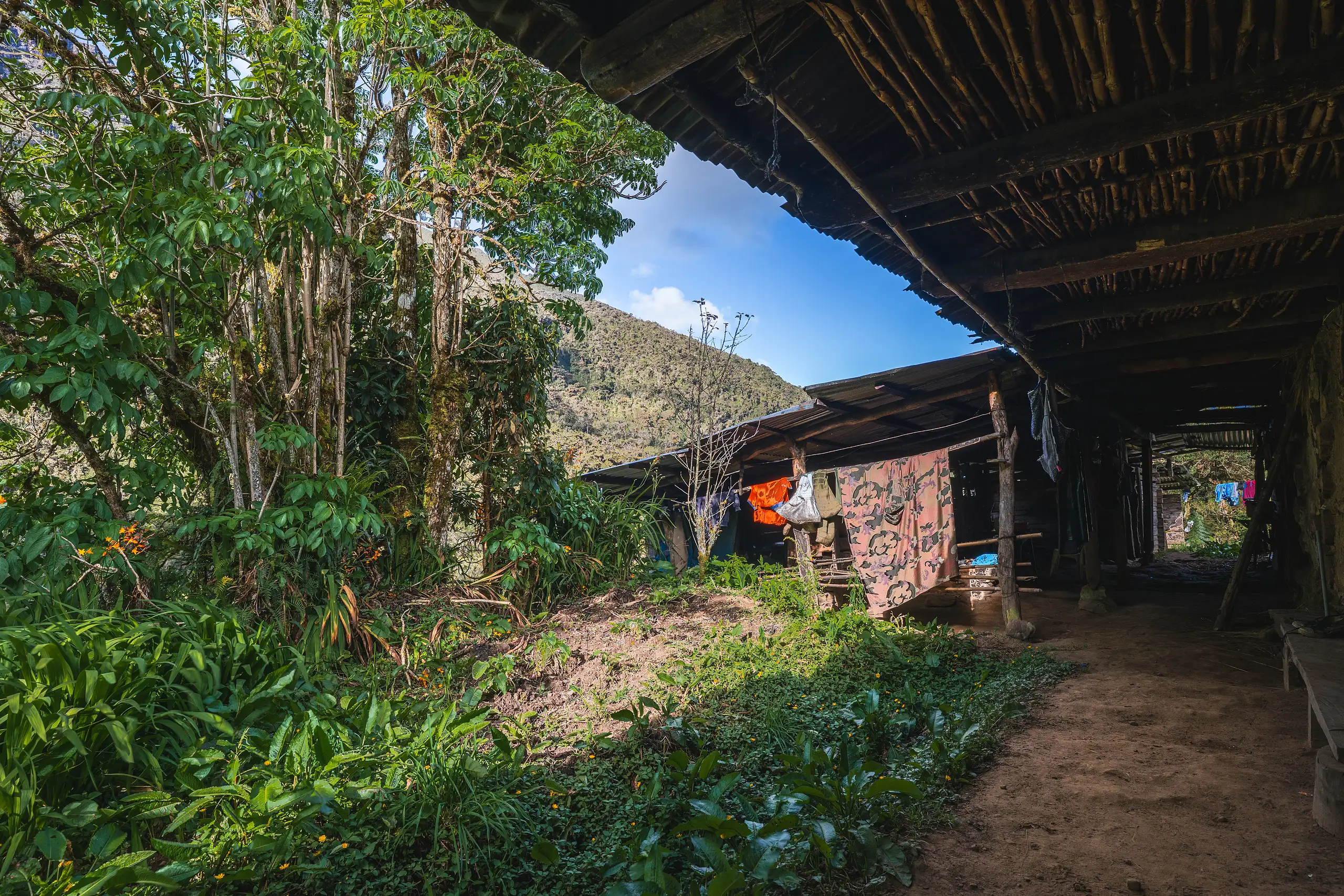 A long wooden cabin veranda under a pole roof, a dirt path running past lush trees and blankets hung to dry, a green hillside and blue sky beyond, Laguna de los Cóndores, Amazonas, Peru