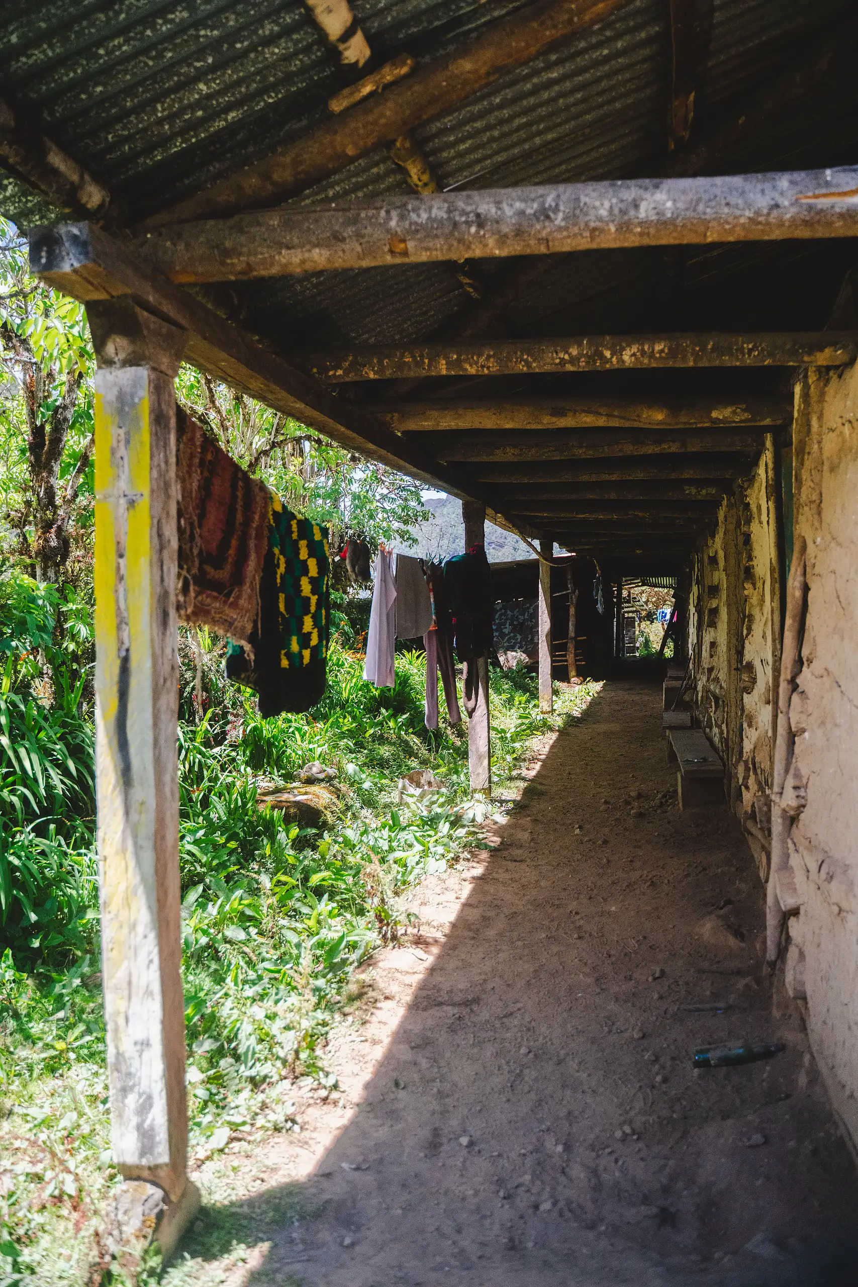 Vertical view down a long covered walkway with a corrugated roof held up by wooden posts, laundry hanging along the side and green plants at the edge, Laguna de los Cóndores, Amazonas, Peru