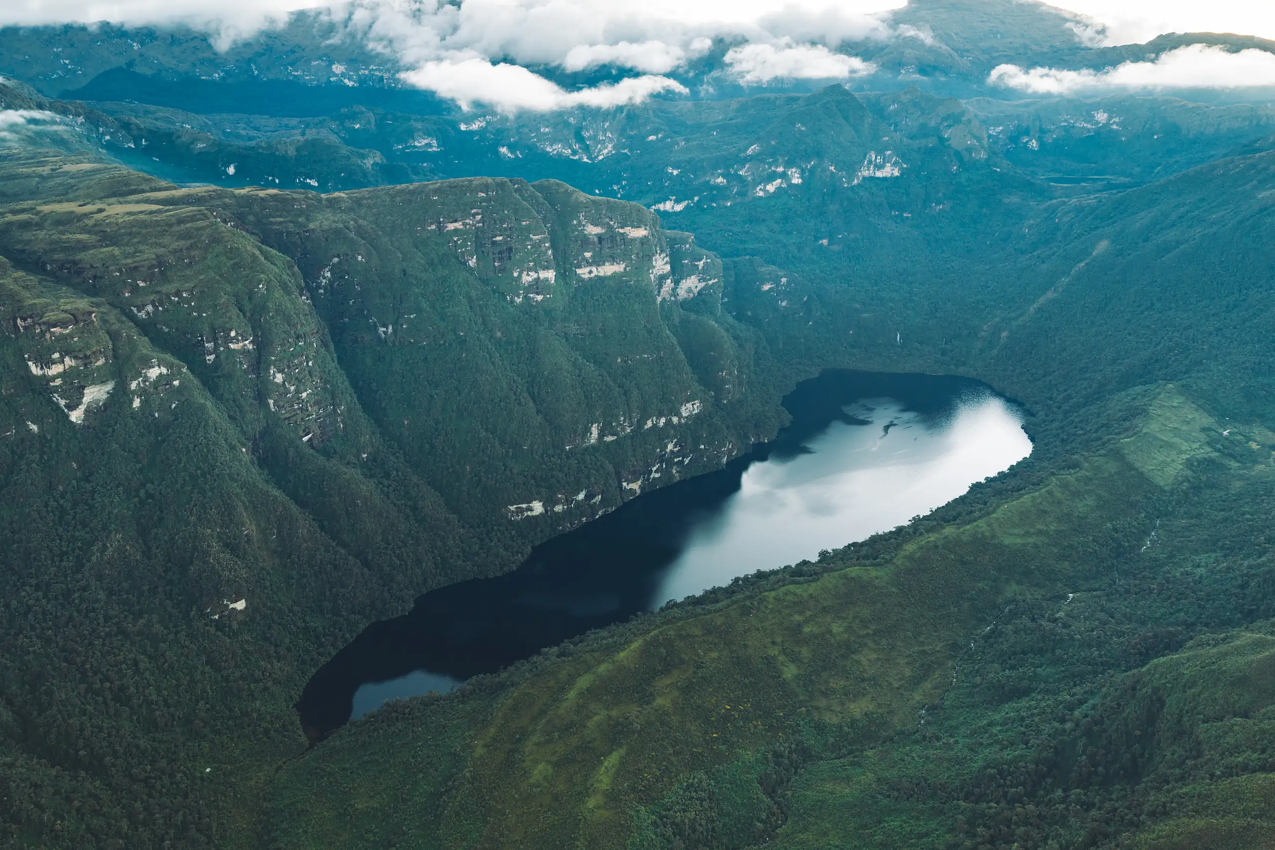 Drone view of a dark lake reflecting white cloud, cupped by steep green slopes and limestone cliffs, more ridges and low cloud beyond, Laguna de los Cóndores, Amazonas, Peru
