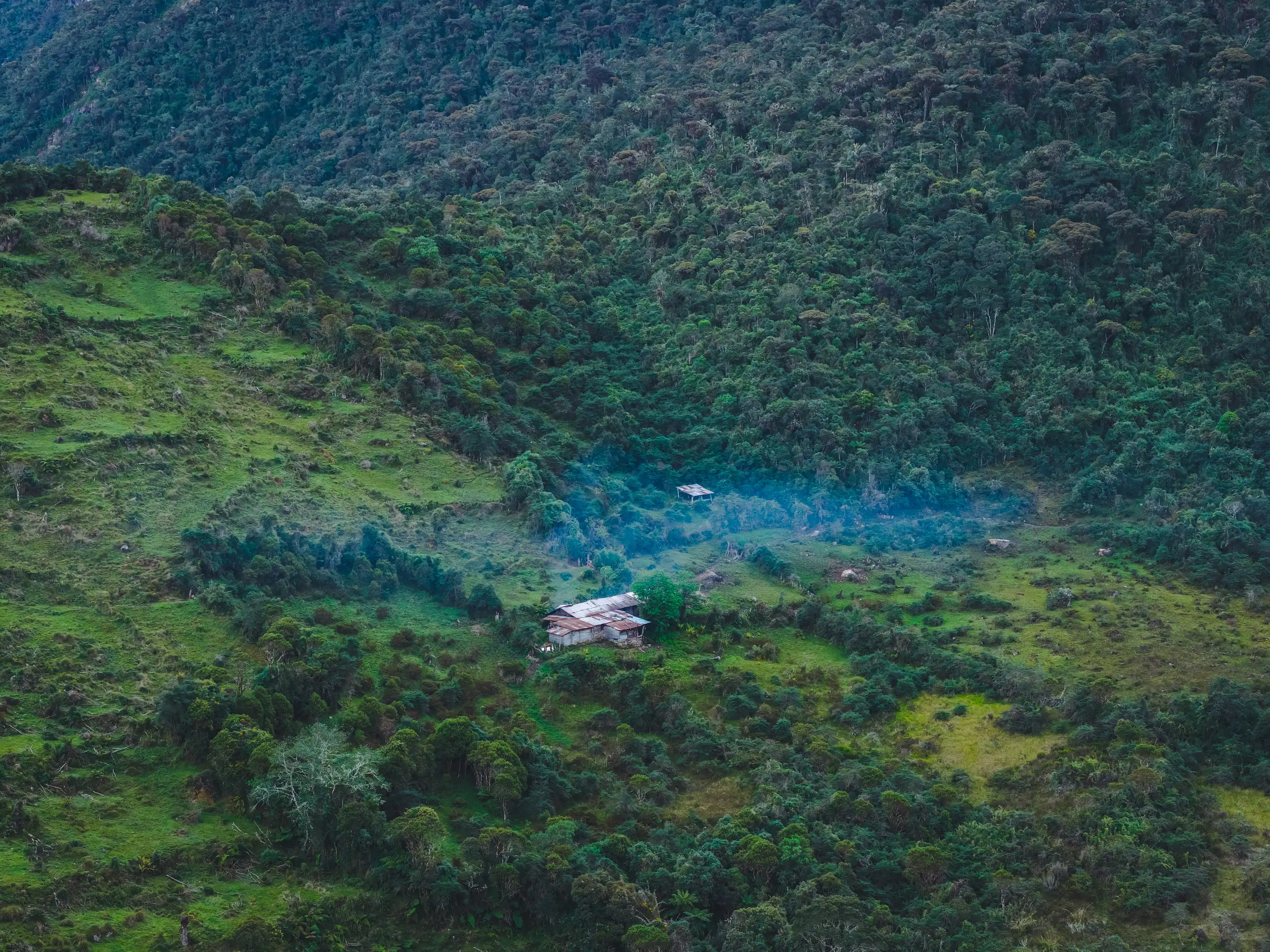 Aerial view of a small tin-roofed cabin in a green clearing, blue woodsmoke drifting across the meadow, cloud forest covering the slopes all around, Laguna de los Cóndores, Amazonas, Peru