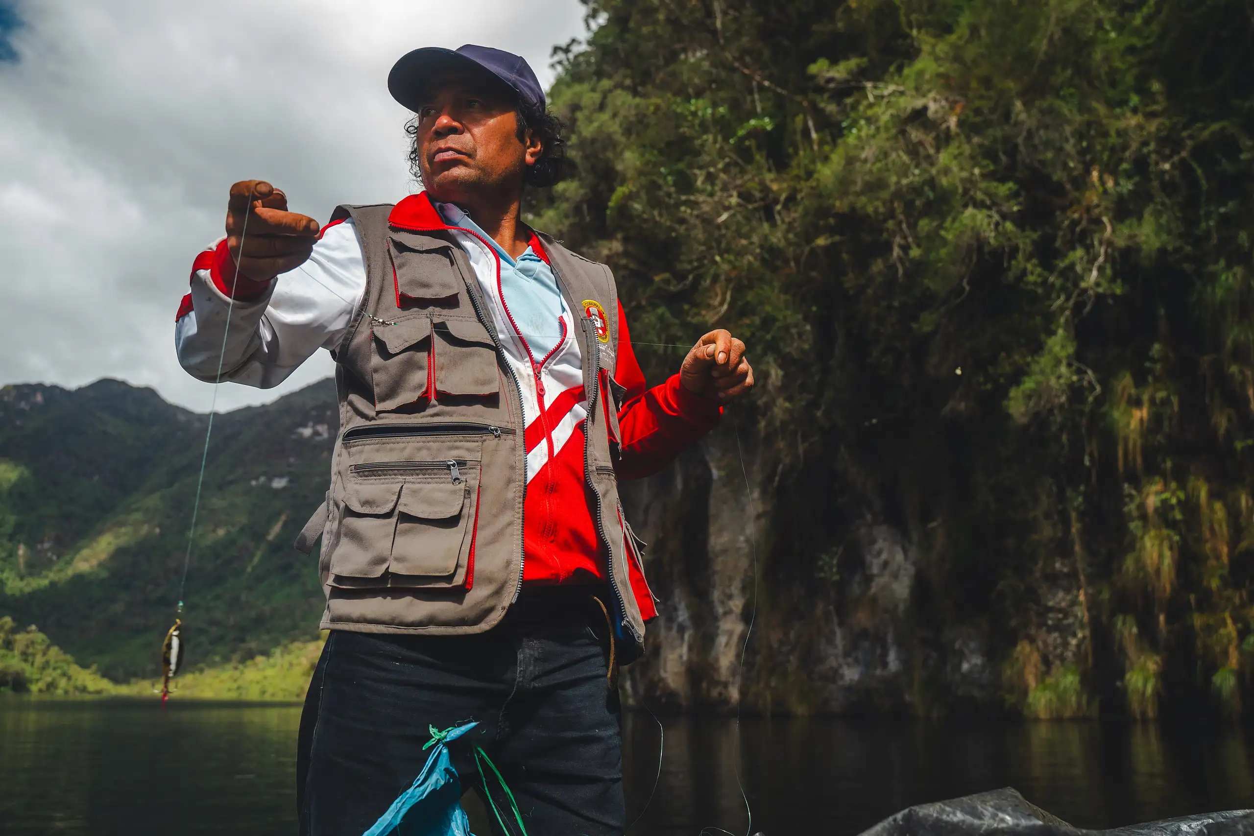 An AAV guide in a cap and a many-pocketed vest holding up a hand line with a small trout on the end, standing in a boat beneath a forested cliff on dark water, Laguna de los Cóndores, Amazonas, Peru