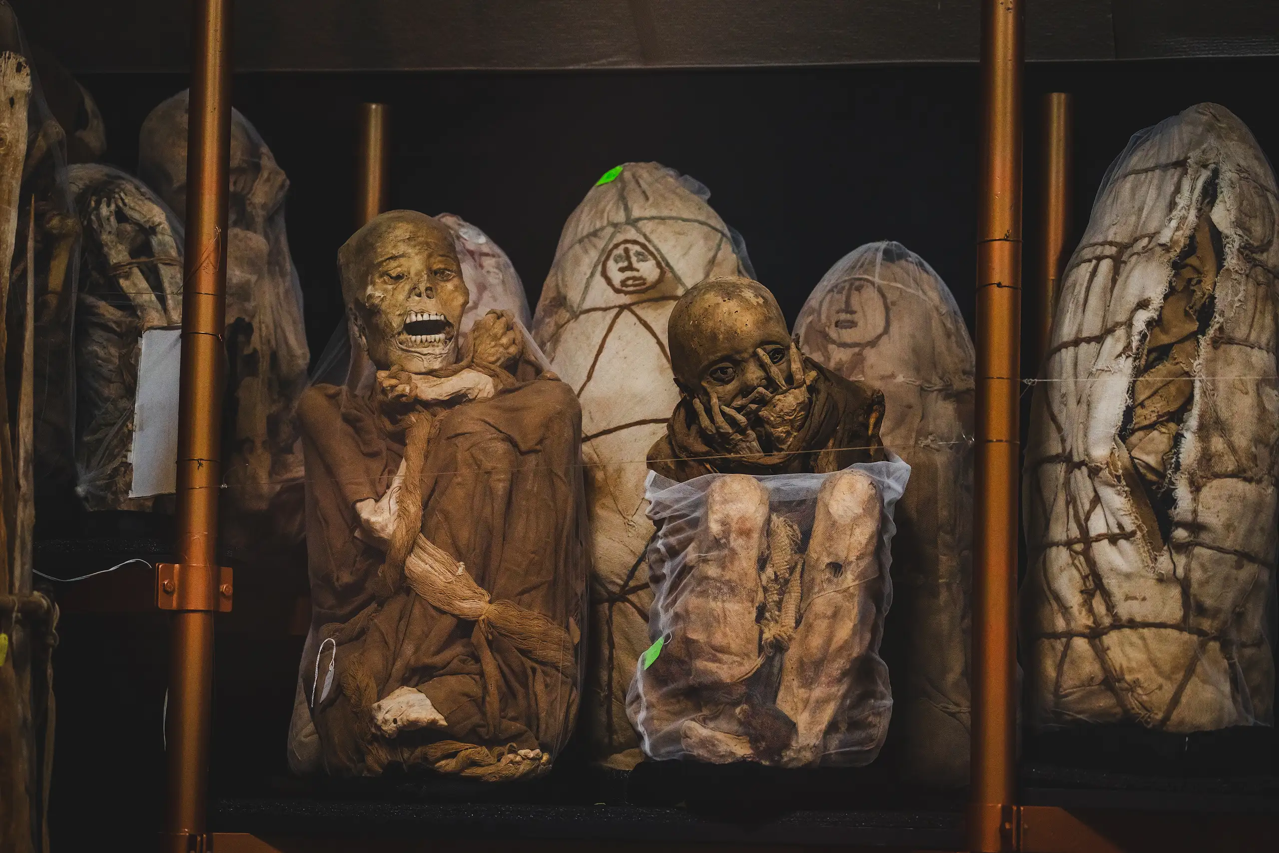 Several seated mummies and cloth funerary bundles, some painted with faces, lined up on shelves between orange metal posts, Museo Leymebamba, Amazonas, Peru