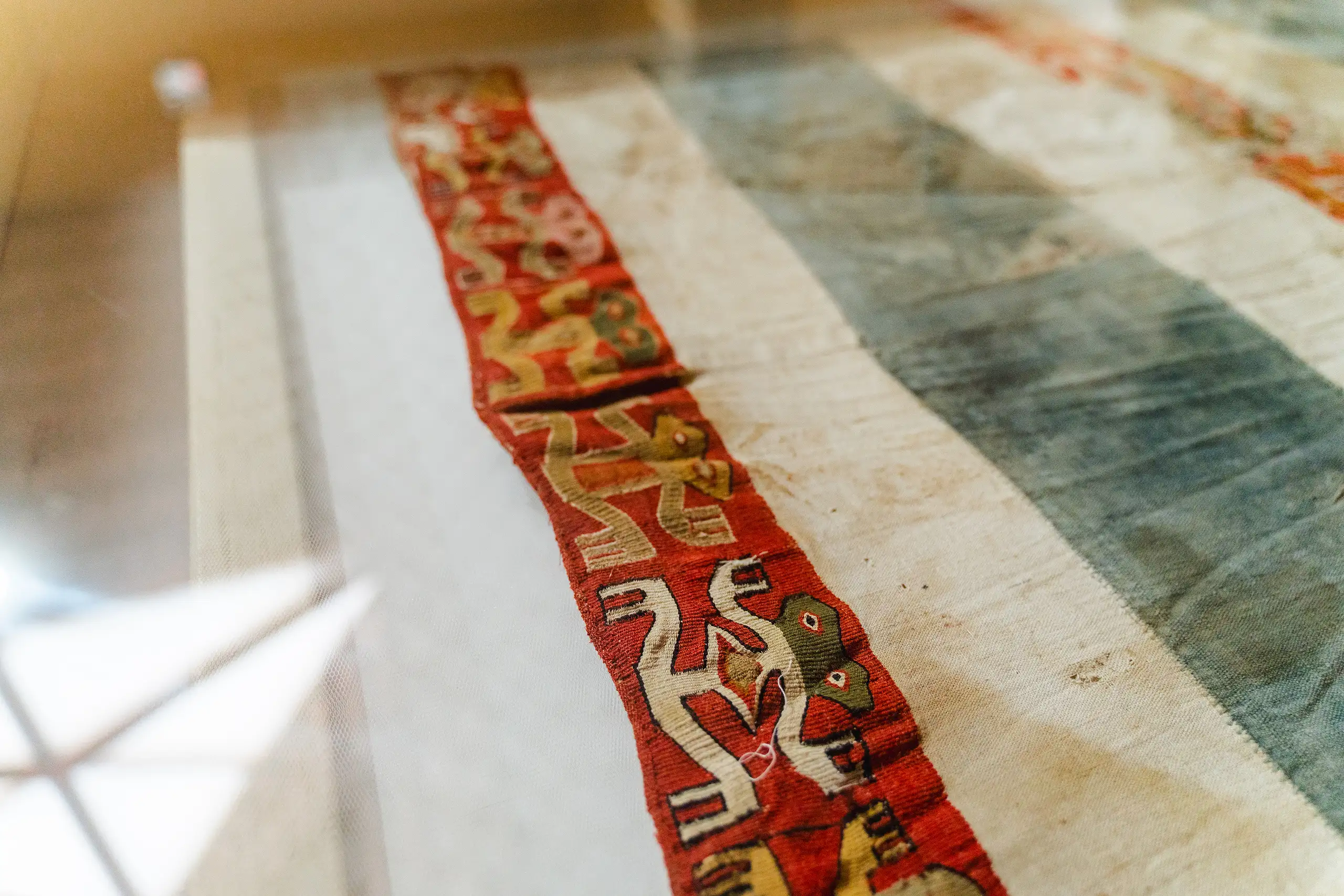 A cream cloth with a woven red band of stylised animal figures in yellow and green, seen through display glass, Museo Leymebamba, Amazonas, Peru