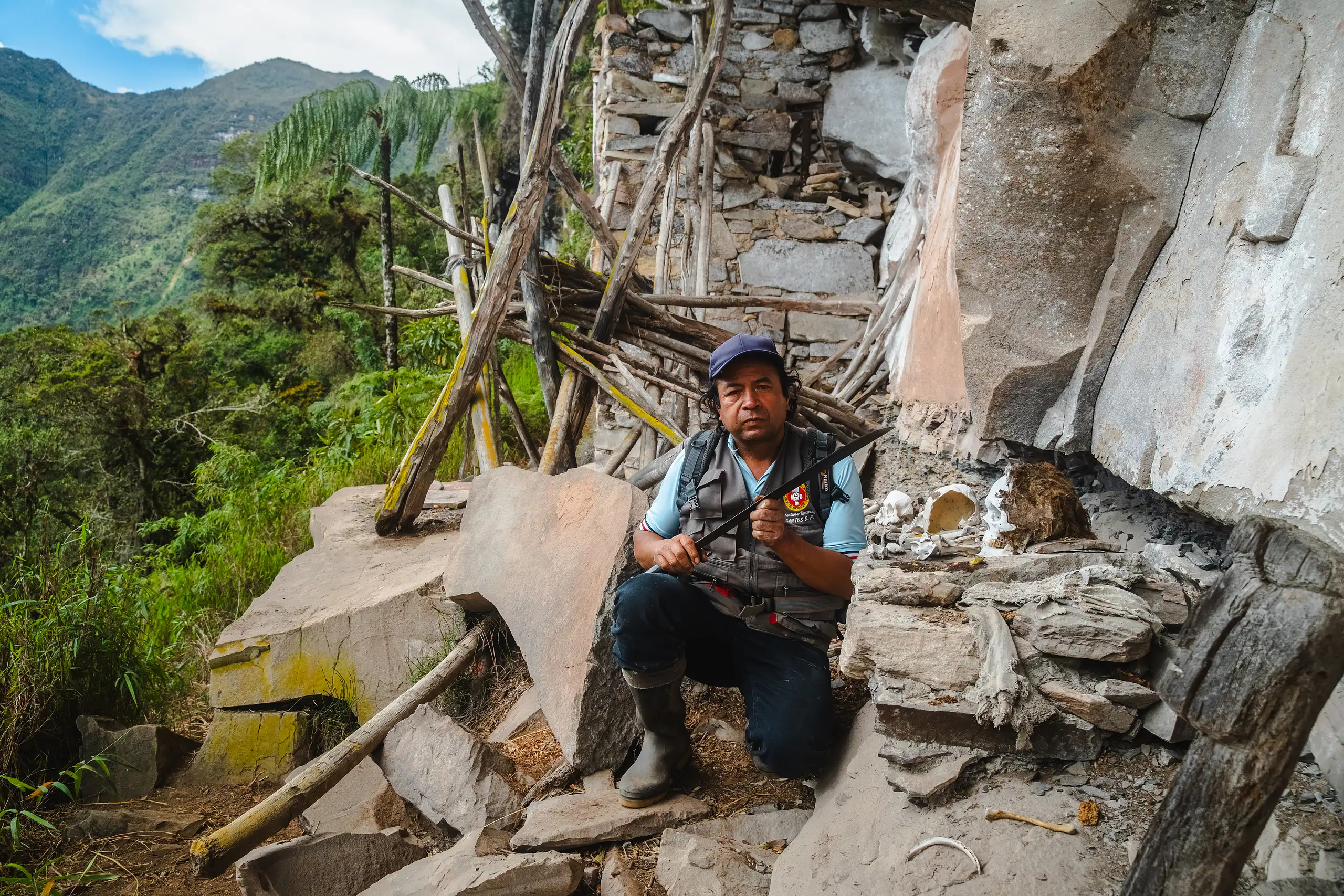 An AAV guide crouching on a rock ledge beside Chachapoya stone funerary walls and a limestone overhang, wooden poles leaning against the ruins and forested mountains behind, Laguna de los Cóndores, Amazonas, Peru