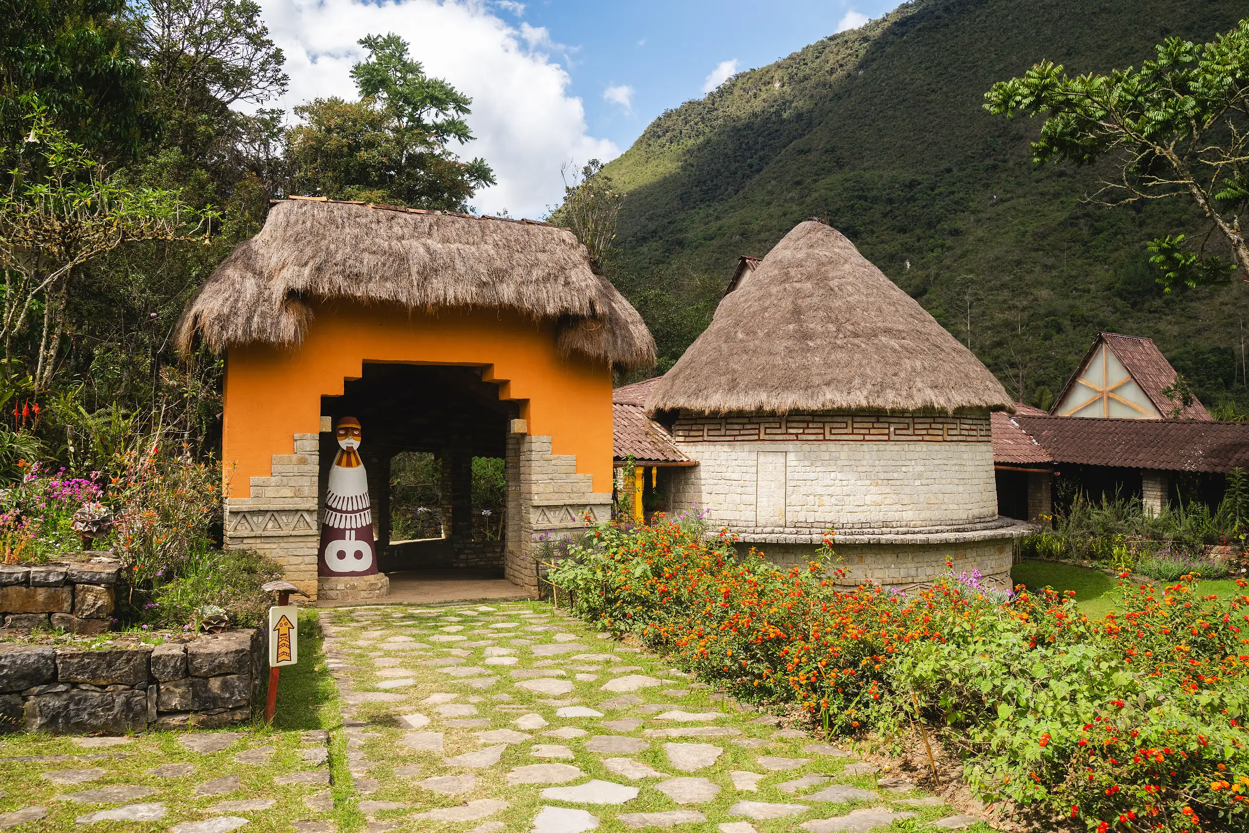 An orange gatehouse with a thatched roof and a round thatched stone building beside it, a stone path lined with red flowers leading in, green mountains behind, Museo Leymebamba, Amazonas, Peru