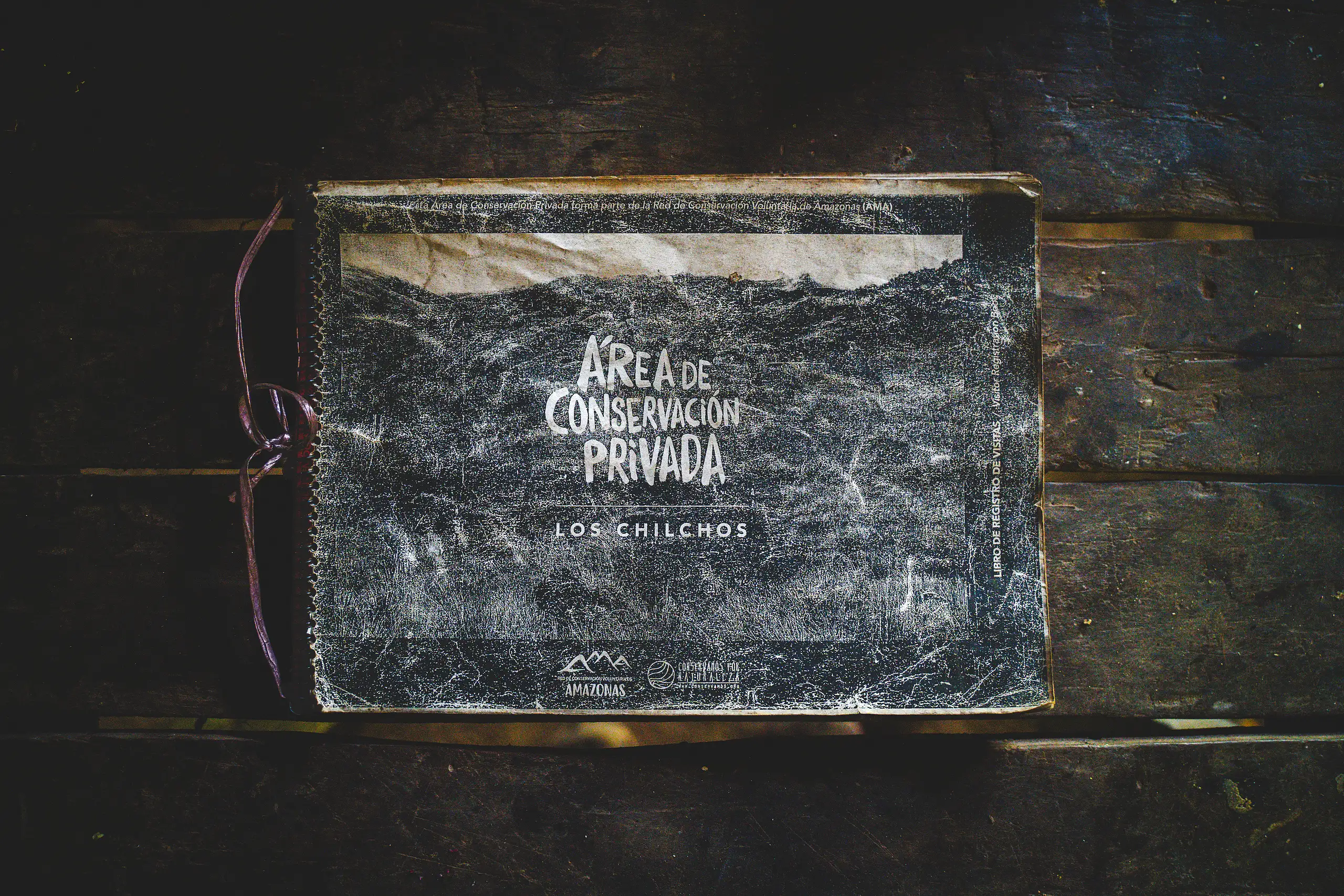 A worn spiral guest log book with a chalk-style cover reading Área de Conservación Privada Los Chilchos, lying on dark wooden boards, Laguna de los Cóndores, Amazonas, Peru
