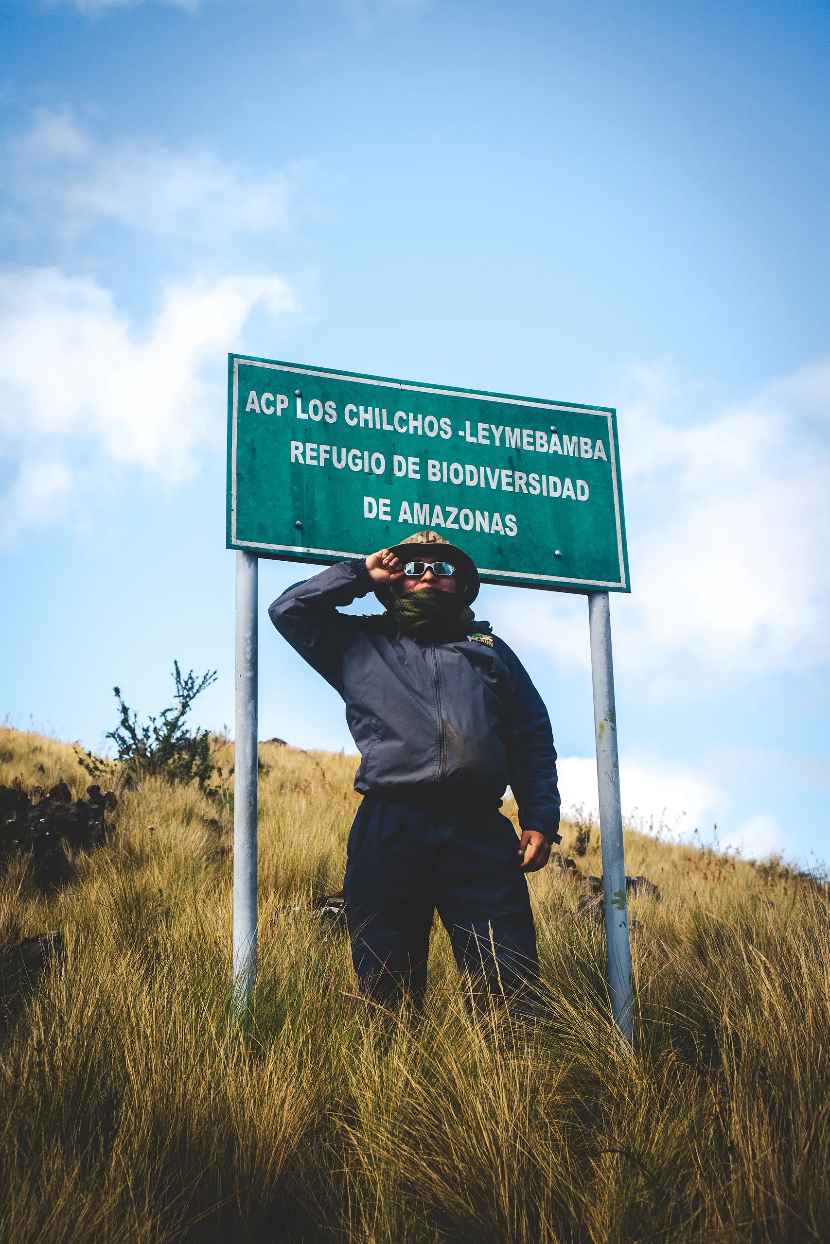 A trekker shading their eyes in tall golden grass beneath a green sign reading ACP Los Chilchos-Leymebamba, Refugio de Biodiversidad de Amazonas, blue sky behind, Laguna de los Cóndores, Amazonas, Peru