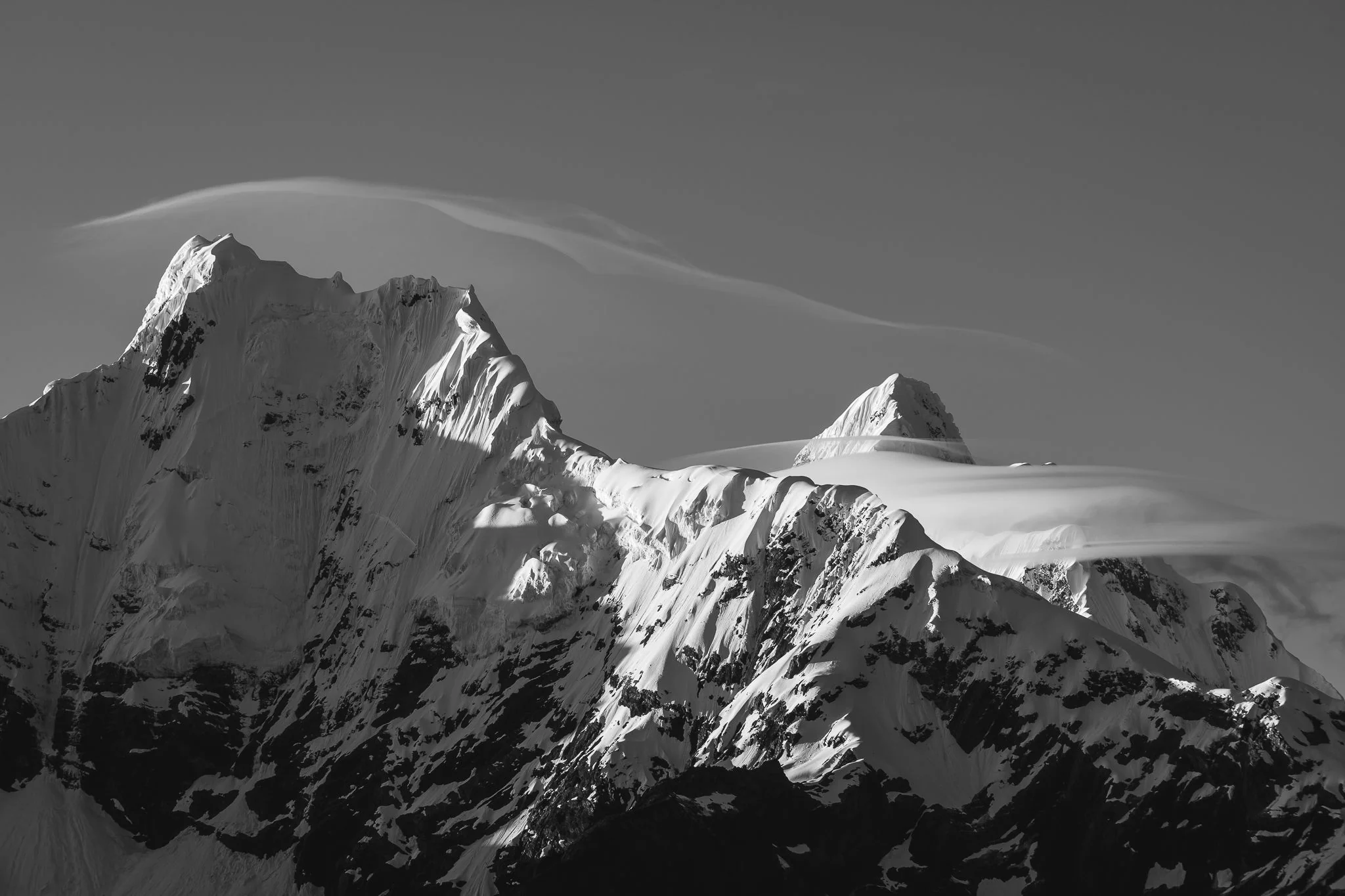 Black-and-white view of the snow and rock summits of Nevado San Juan with lens-shaped clouds above them, from the Quilcayhuanca valley, Peru