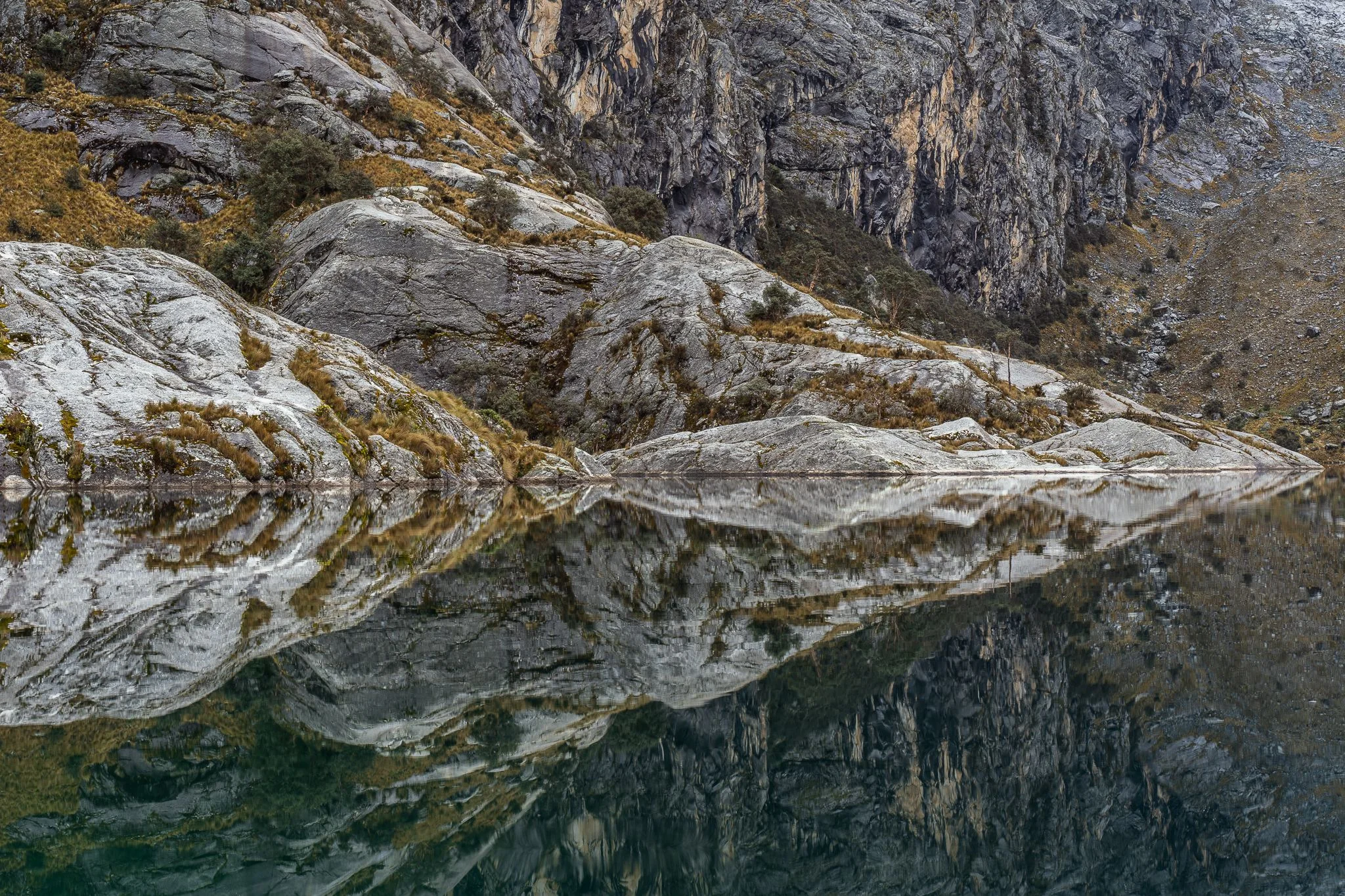 Pale rock slabs and grass on the shoreline mirrored perfectly in the still green water of Laguna Churup, Cordillera Blanca, Peru