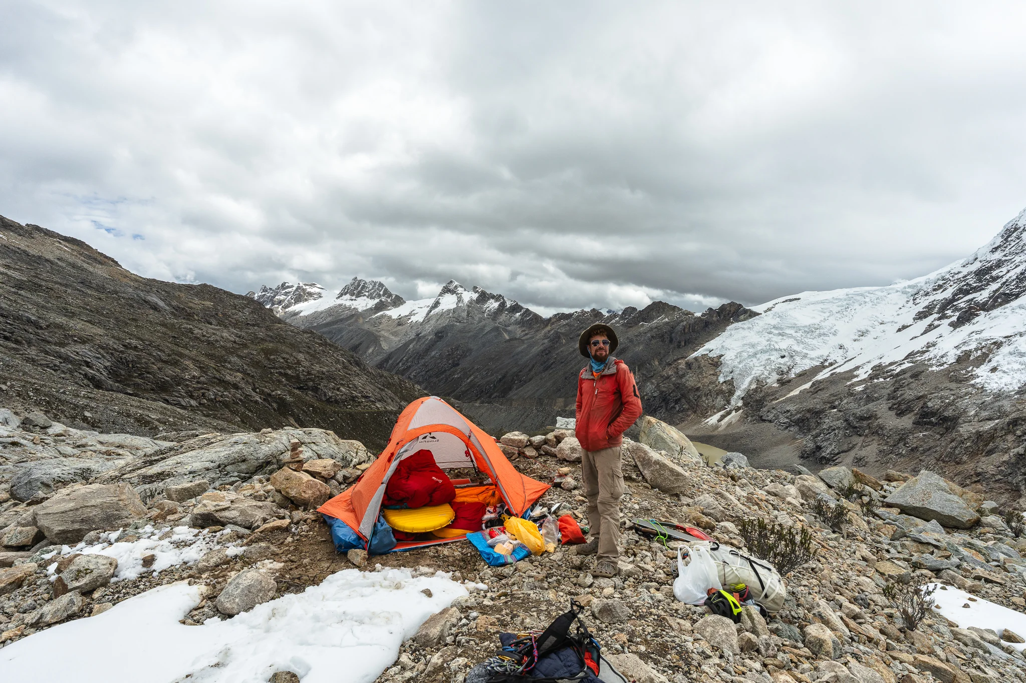 A climber standing beside an orange tent at a rocky moraine camp, gear spread on the ground and a glacier spilling from the peaks behind, below Nevado Palcaraju, Peru