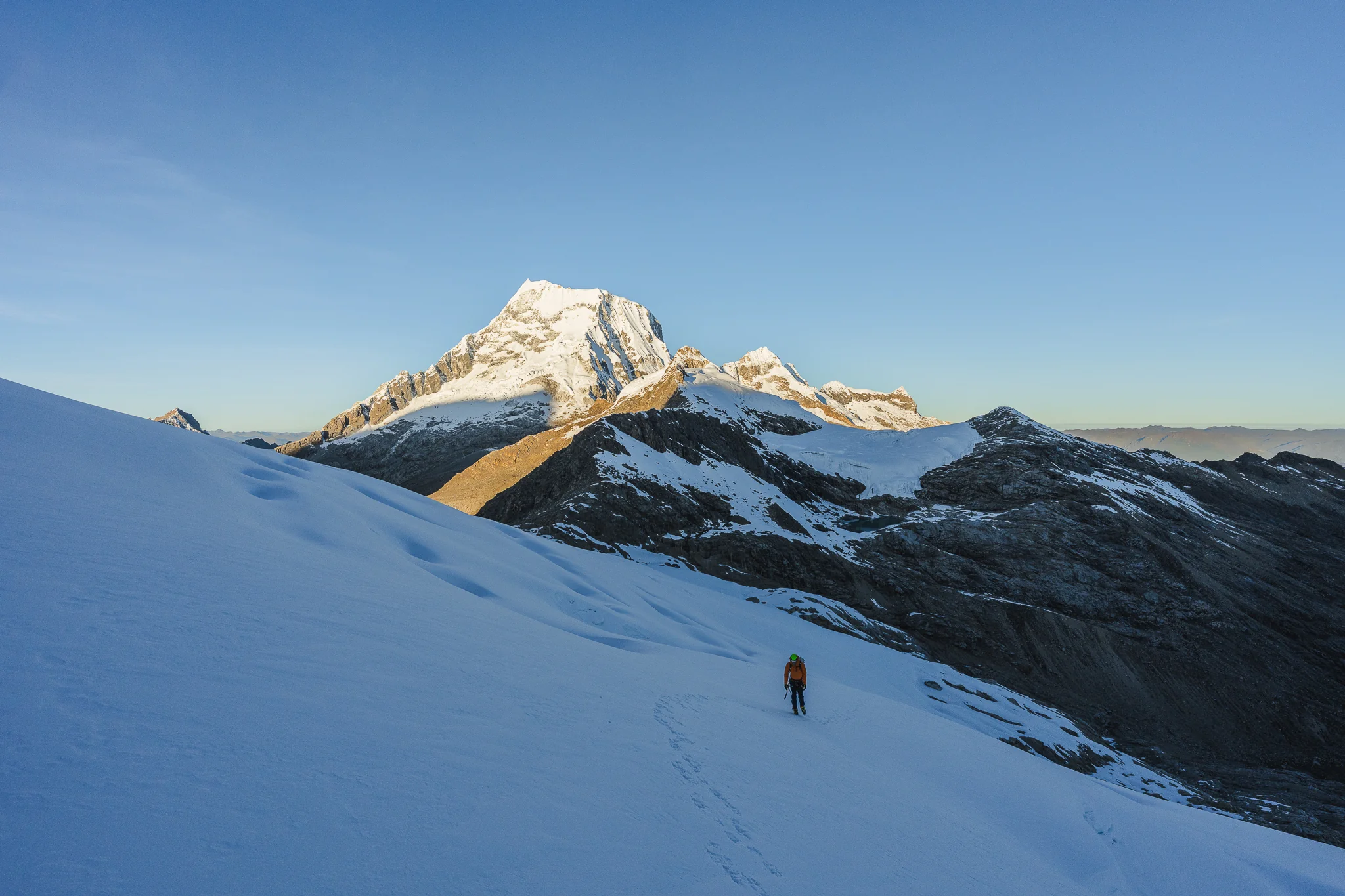A climber crossing a broad snowfield at sunrise towards the sunlit summit of Nevado Ranrapalca, on the approach to high camp on Palcaraju, Peru
