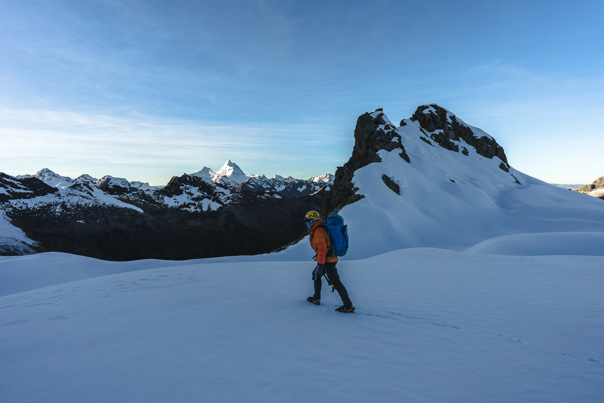 A climber with a large pack walking a snow slope in cold morning light, the pyramid of a distant snow peak on the skyline, below Nevado Palcaraju West, Peru