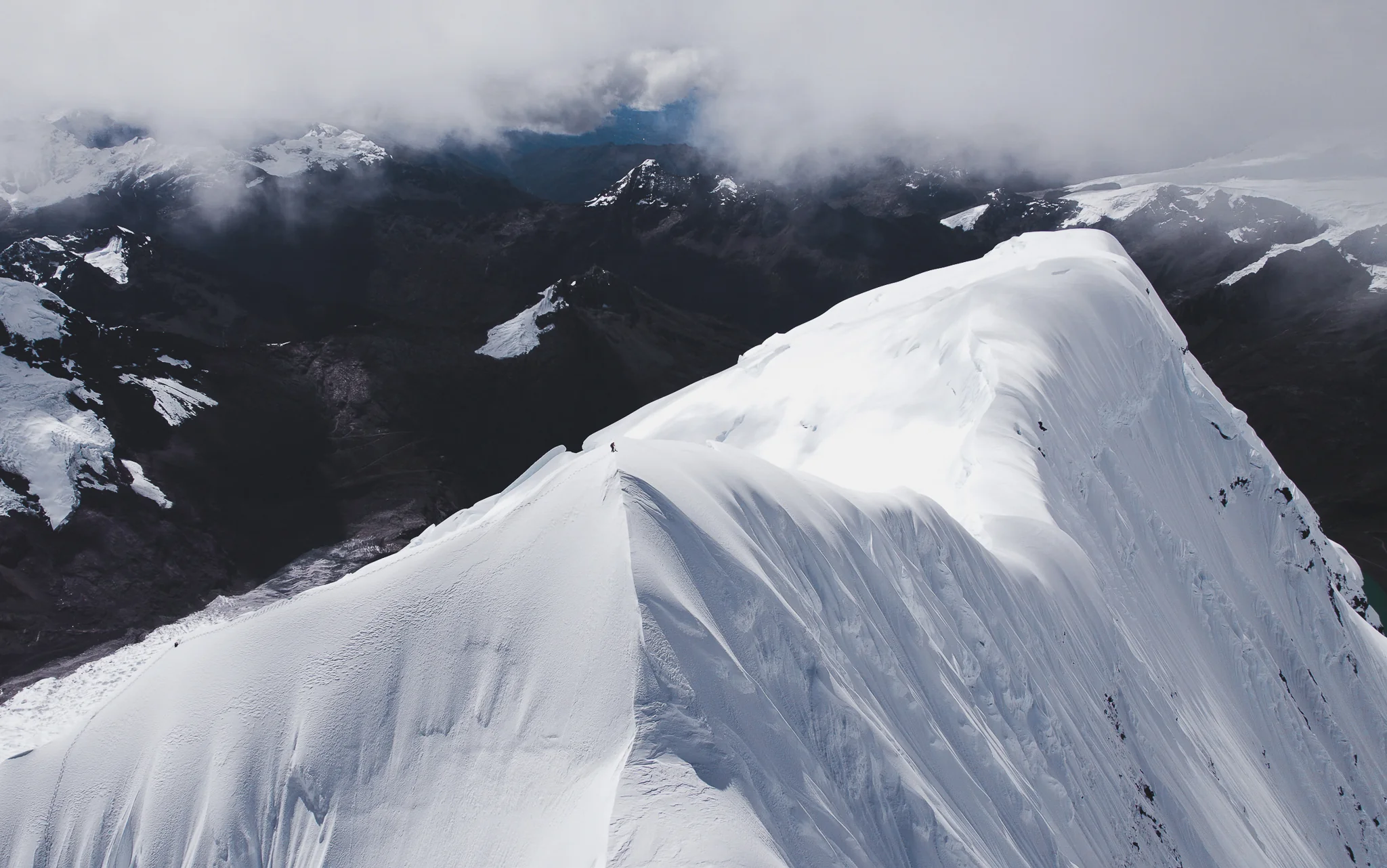 Nate Heald, a tiny figure on the broad corniced snow summit of Nevado Palcaraju, above dark peaks partly hidden in cloud, Cordillera Blanca, Peru