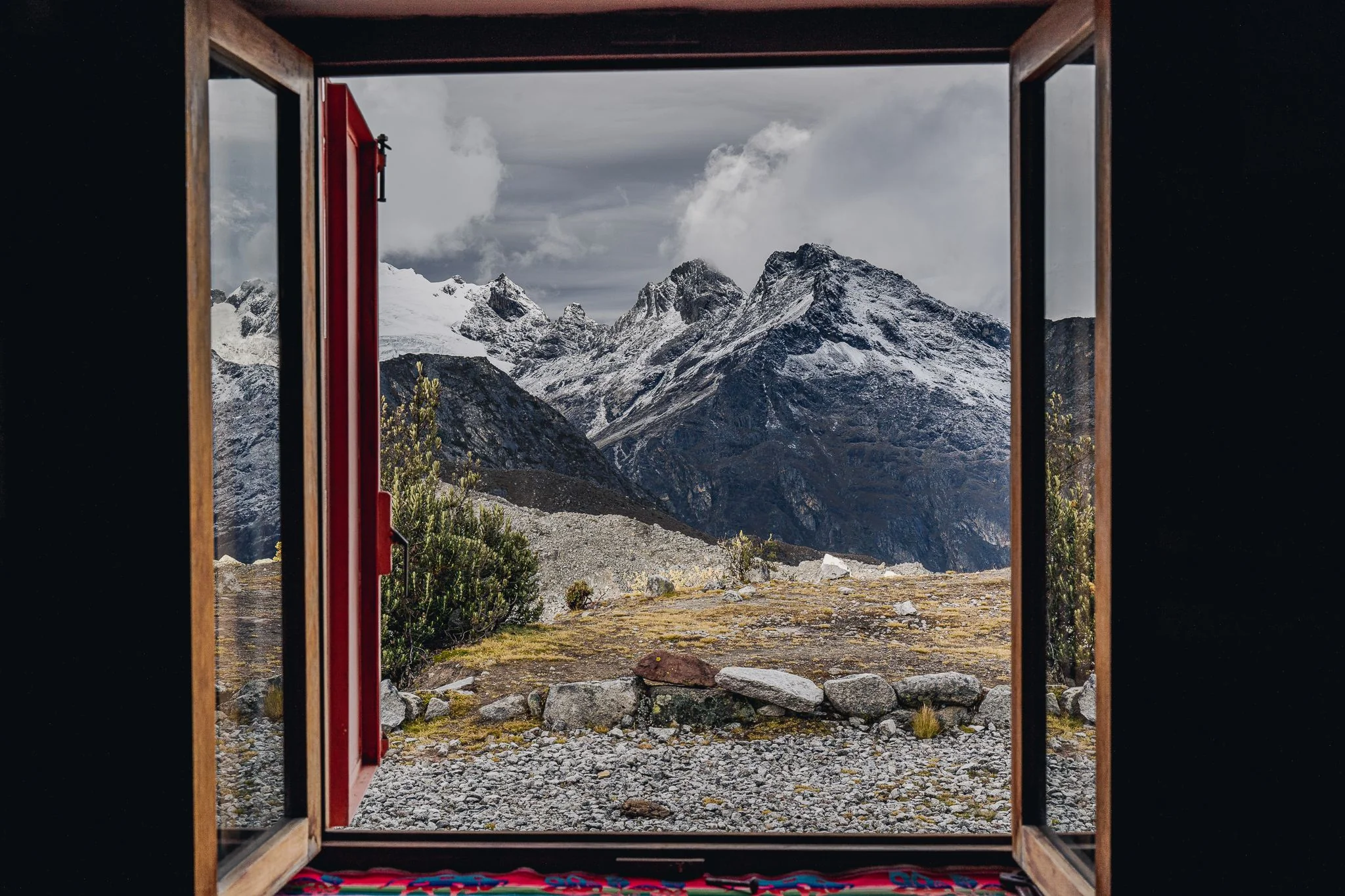 Snow-covered peaks framed in the open wooden window of Refugio Pisco, a woven blanket on the sill, Cordillera Blanca, Peru