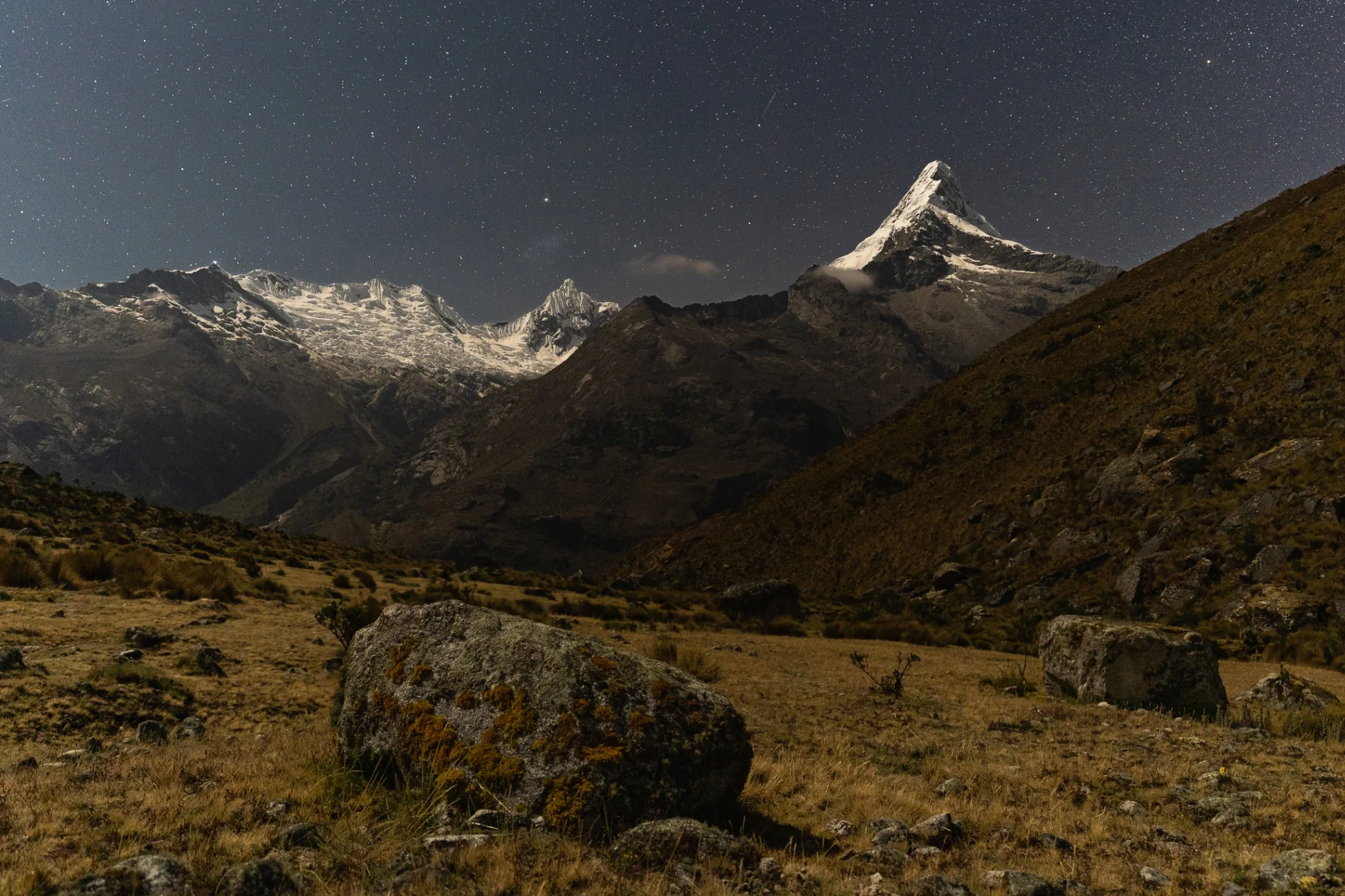 The pyramid of Nevado Artesonraju lit by moonlight under a sky full of stars, boulders on the dark grassland of Alpamayo base camp in front, Peru
