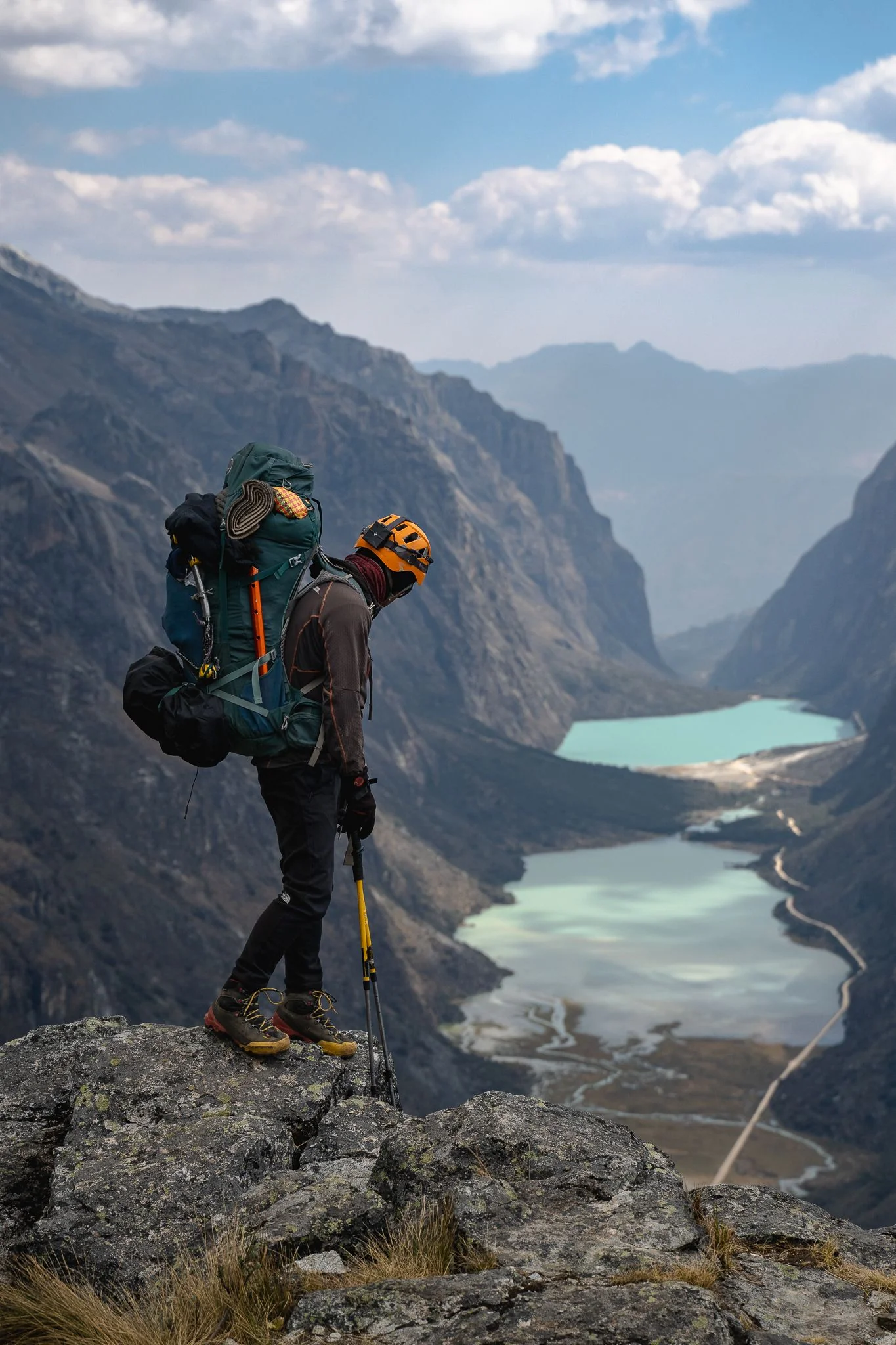 A climber with a helmet, large backpack and ice axe on a rock ledge at the Yanapaccha trailhead, looking down the Llanganuco valley and its two turquoise lakes, Cordillera Blanca, Peru