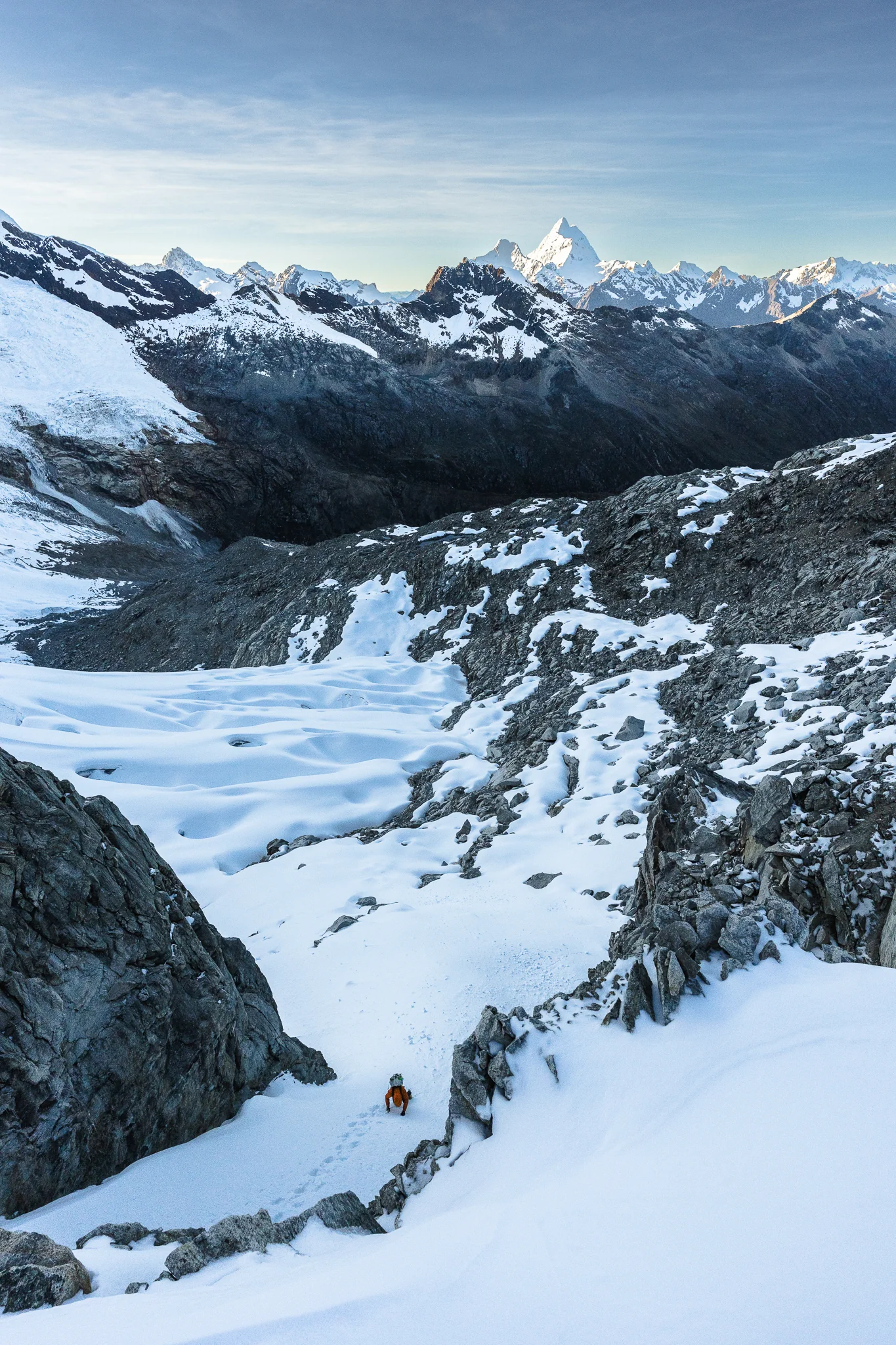 A climber descending a narrow snow chute below Nevado Palcaraju West at dawn, the distant pyramid of Nevado Huantsán catching first light, Peru