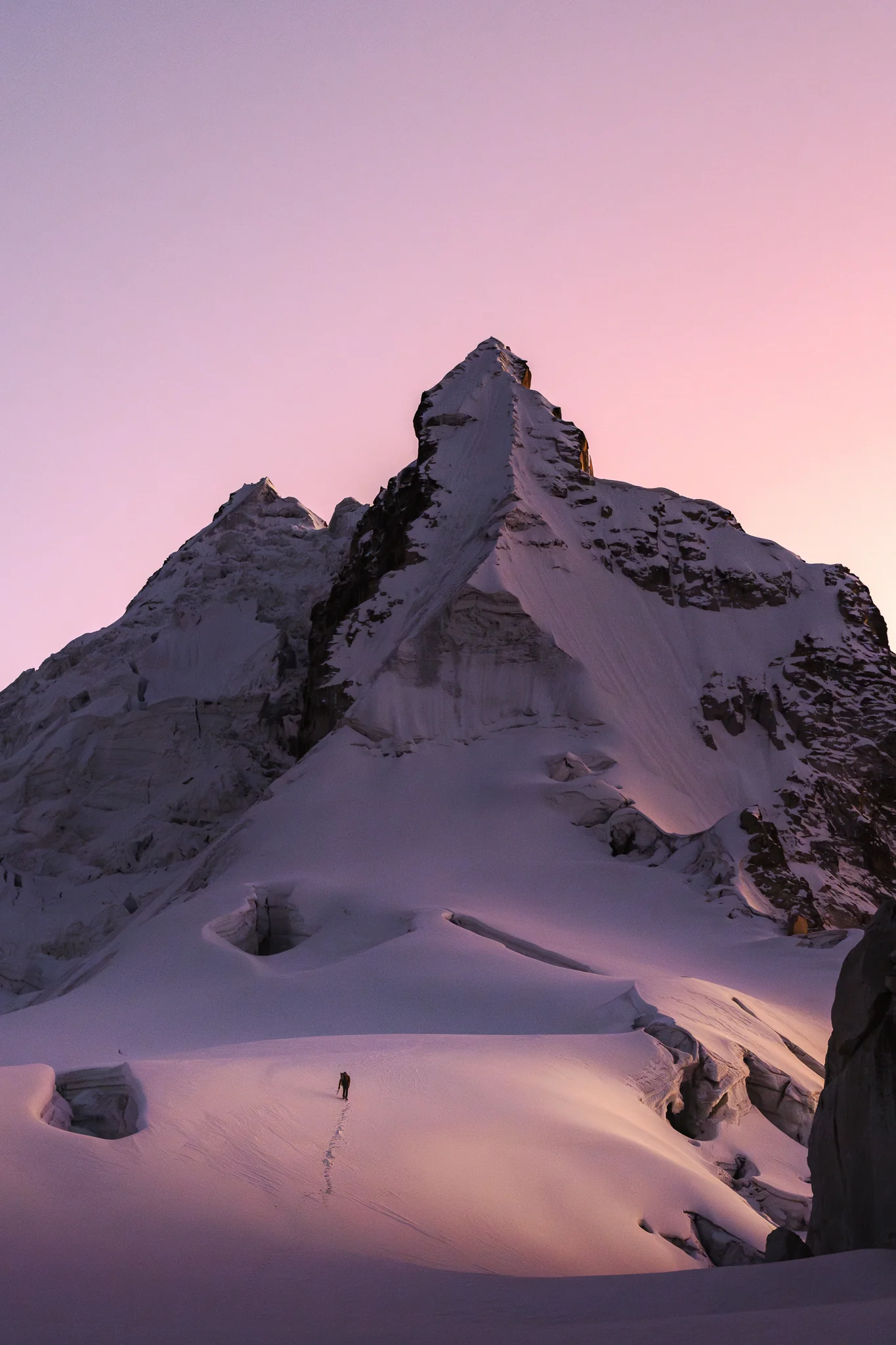 A lone climber crossing a snowfield under the fluted snow ridges of the south peak of Nevado Shaqsha, the sky pink before sunrise, Peru