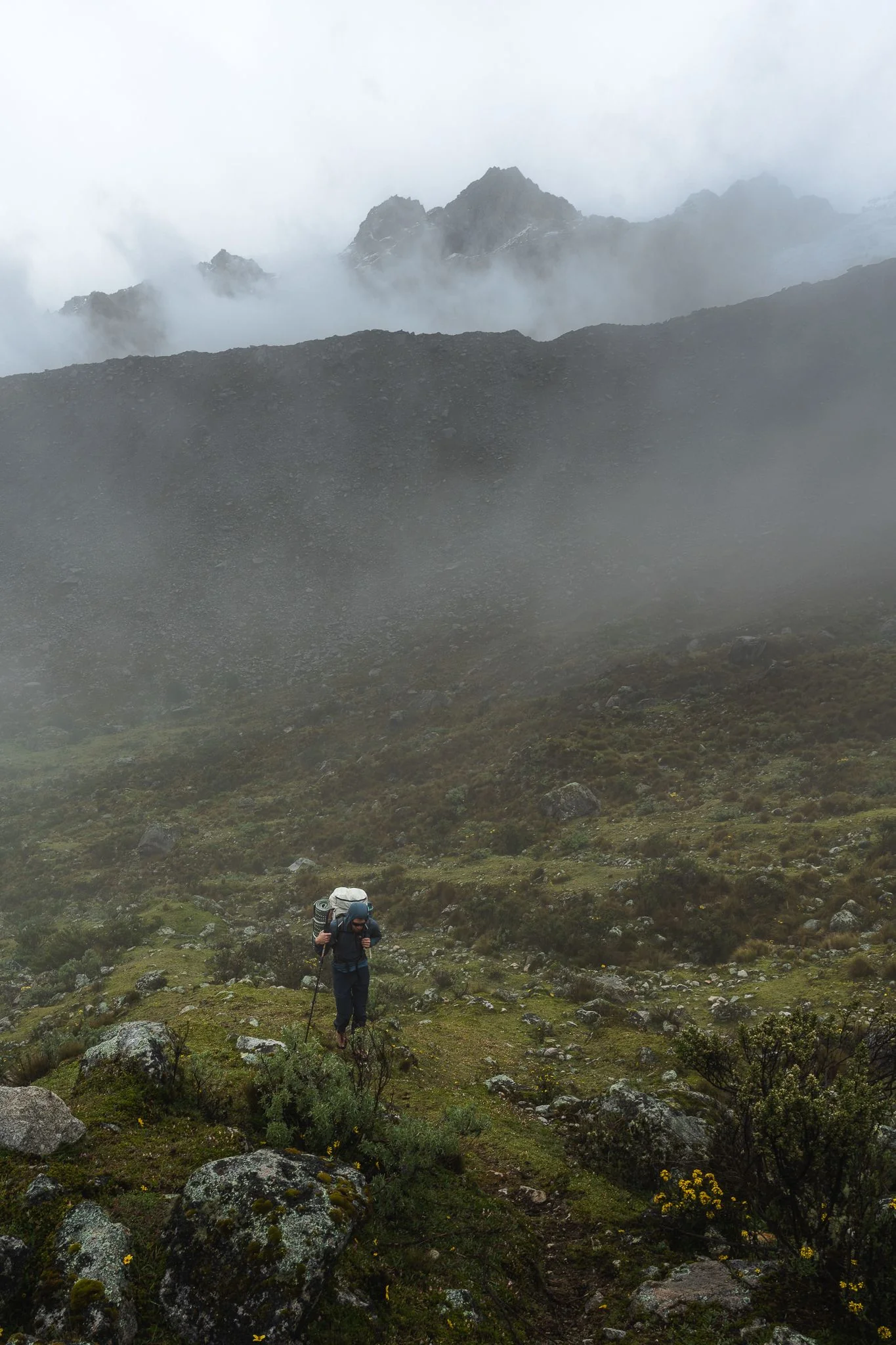 A climber with a large pack walking up a green slope as cloud closes in around the ridges above, approaching moraine camp on Nevado Palcaraju, Peru