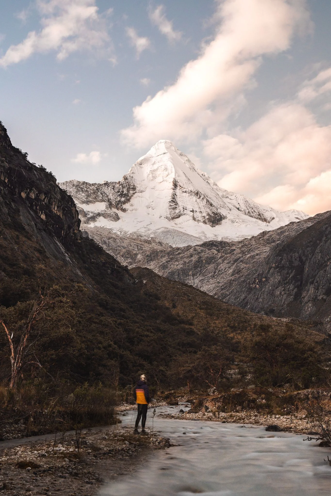 A person standing on a braided riverbed looking up at the snow pyramid of Nevado Artesonraju, dry valley walls on either side, east end of Laguna Parón, Peru