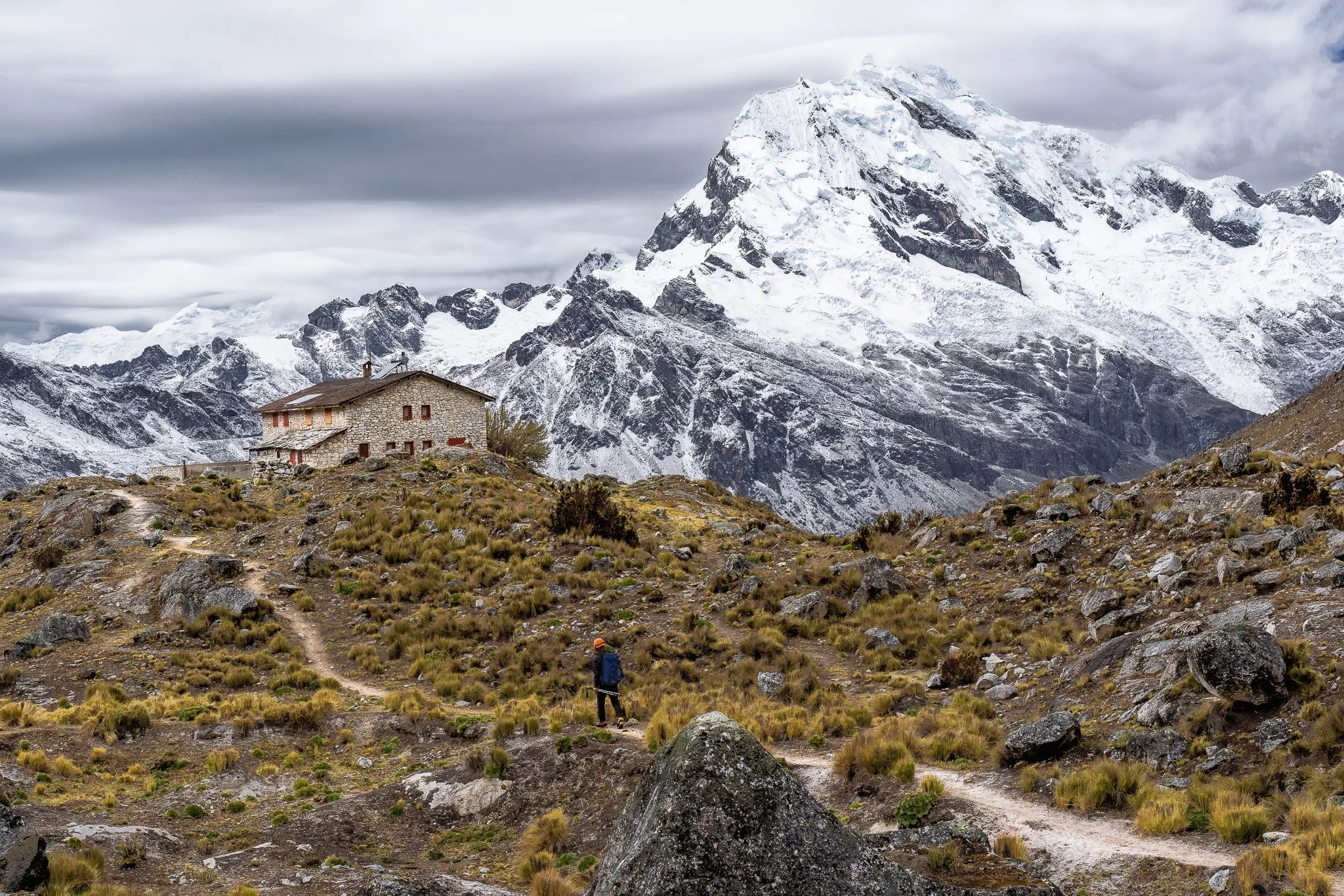 A hiker on a dirt trail through rocky grassland below a stone refuge and a large snow-covered mountain under grey cloud, Cordillera Blanca, Peru