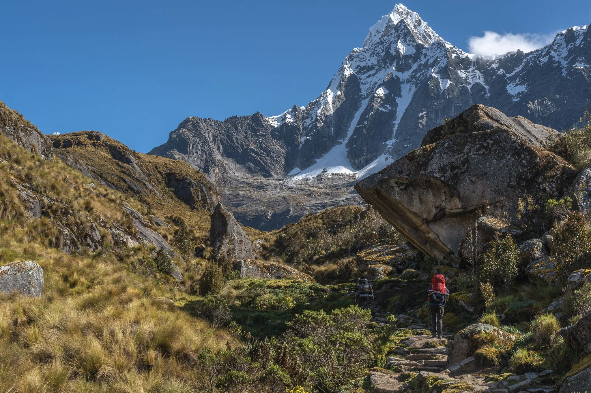A trekker on the trail passing beneath a huge boulder, a sharp snow peak rising at the head of the valley, Santa Cruz trek, Cordillera Blanca, Peru