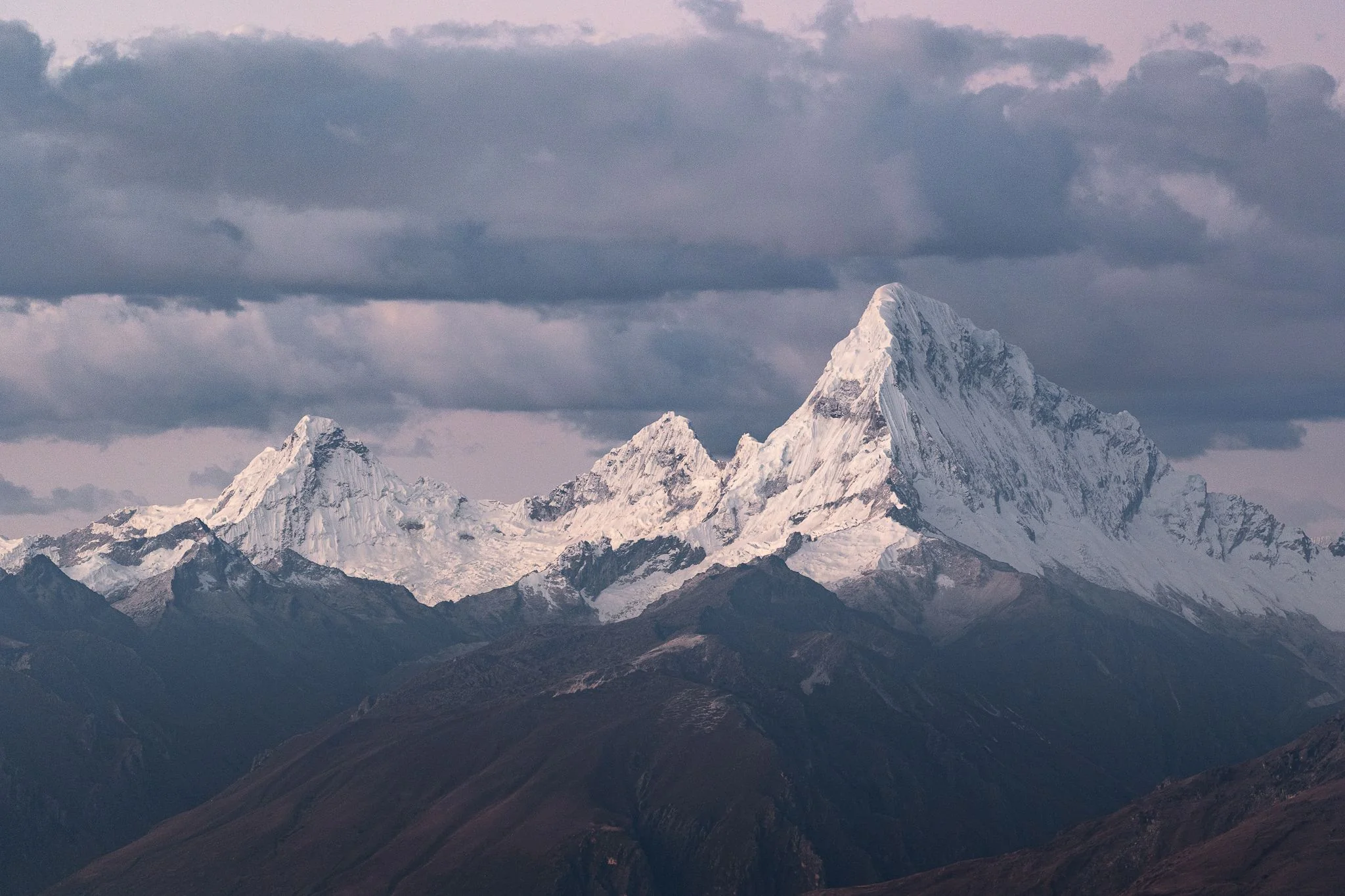 The snow and ice summits of Nevado Santa Cruz Grande under heavy cloud at dusk, seen across dark ridges from the Cordillera Negra, Peru