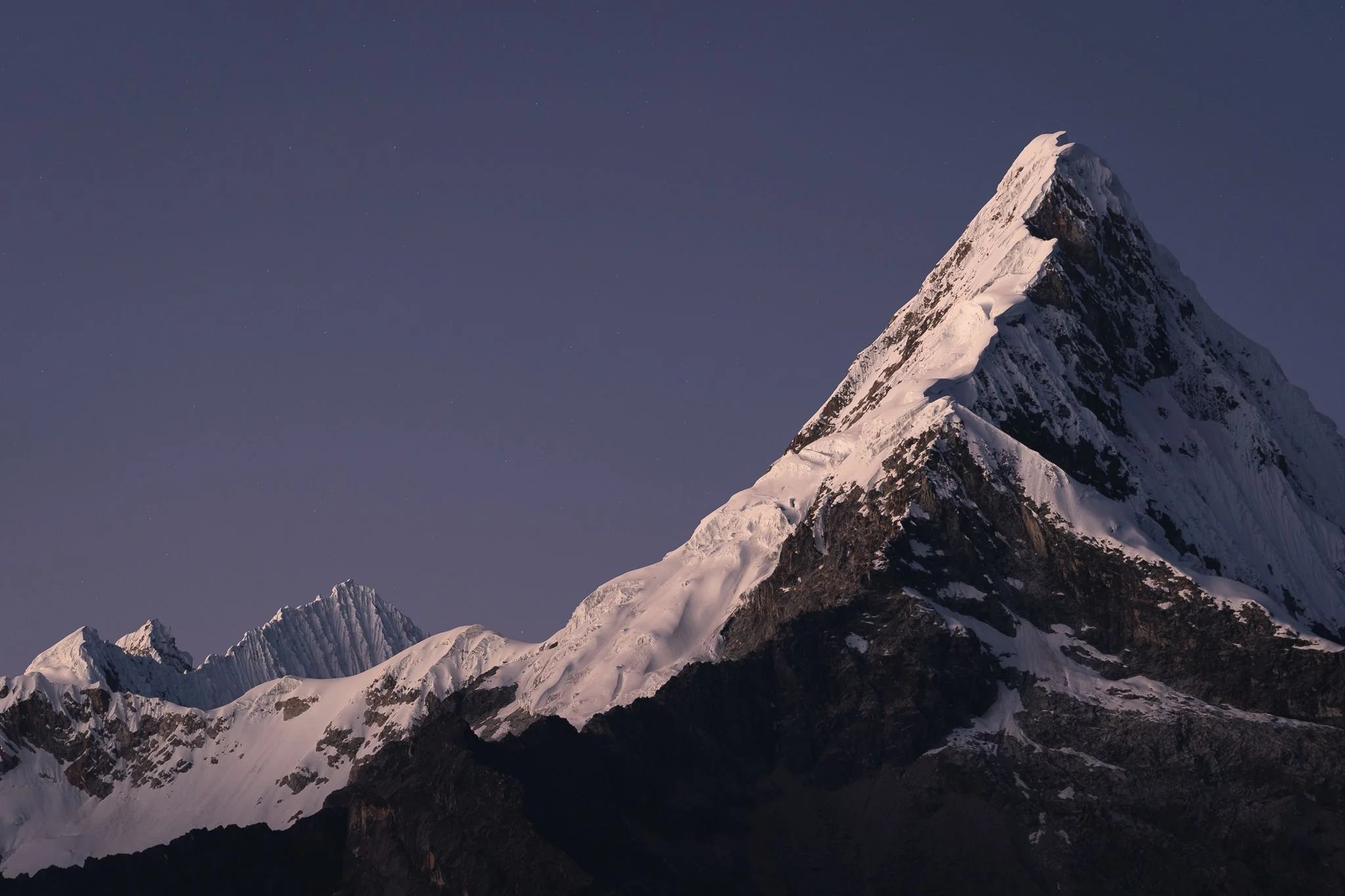 The steep, pyramid-shaped snow peak of Nevado Artesonraju lit pink at dawn against a violet sky, with a jagged snowy ridge beyond, Cordillera Blanca, Peru