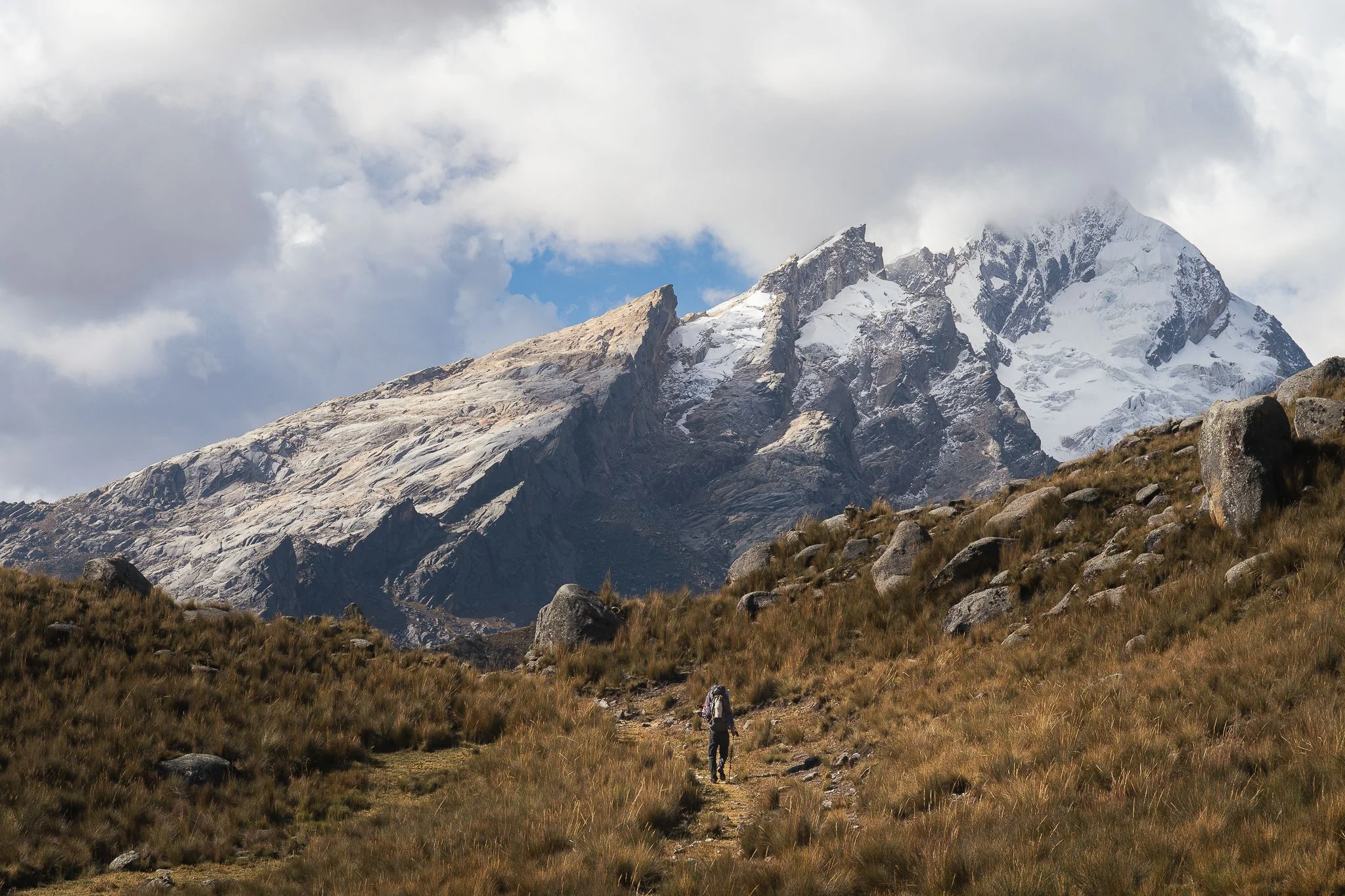 A trekker on a faint path through tall grass below the rock and snow summit of Nevado Shaqsha, cloud building above, Cordillera Blanca, Peru