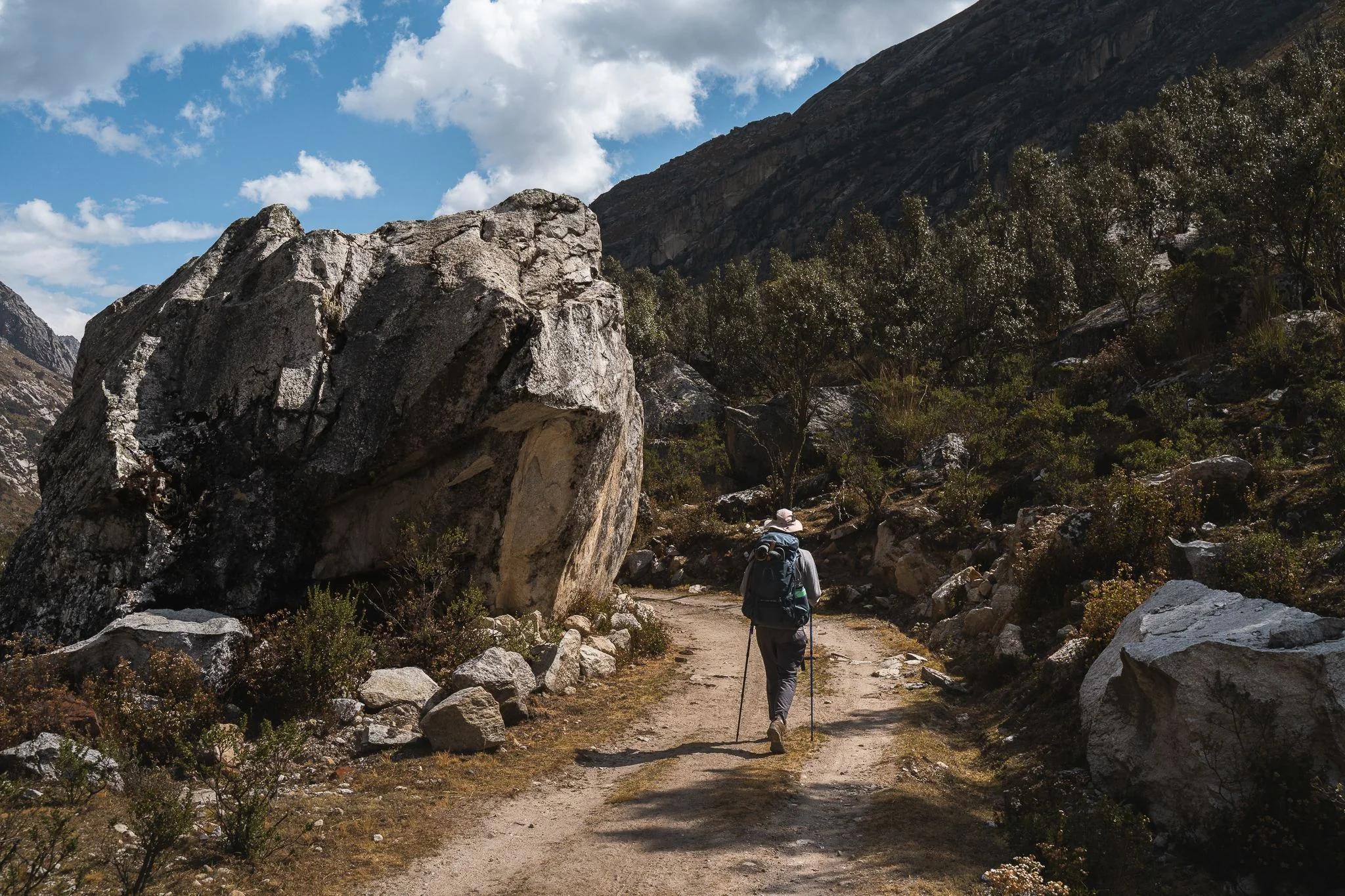 A trekker with walking poles on a dirt trail passing a house-sized boulder, valley walls rising on either side, Quilcayhuanca to Cojup trek, Cordillera Blanca, Peru