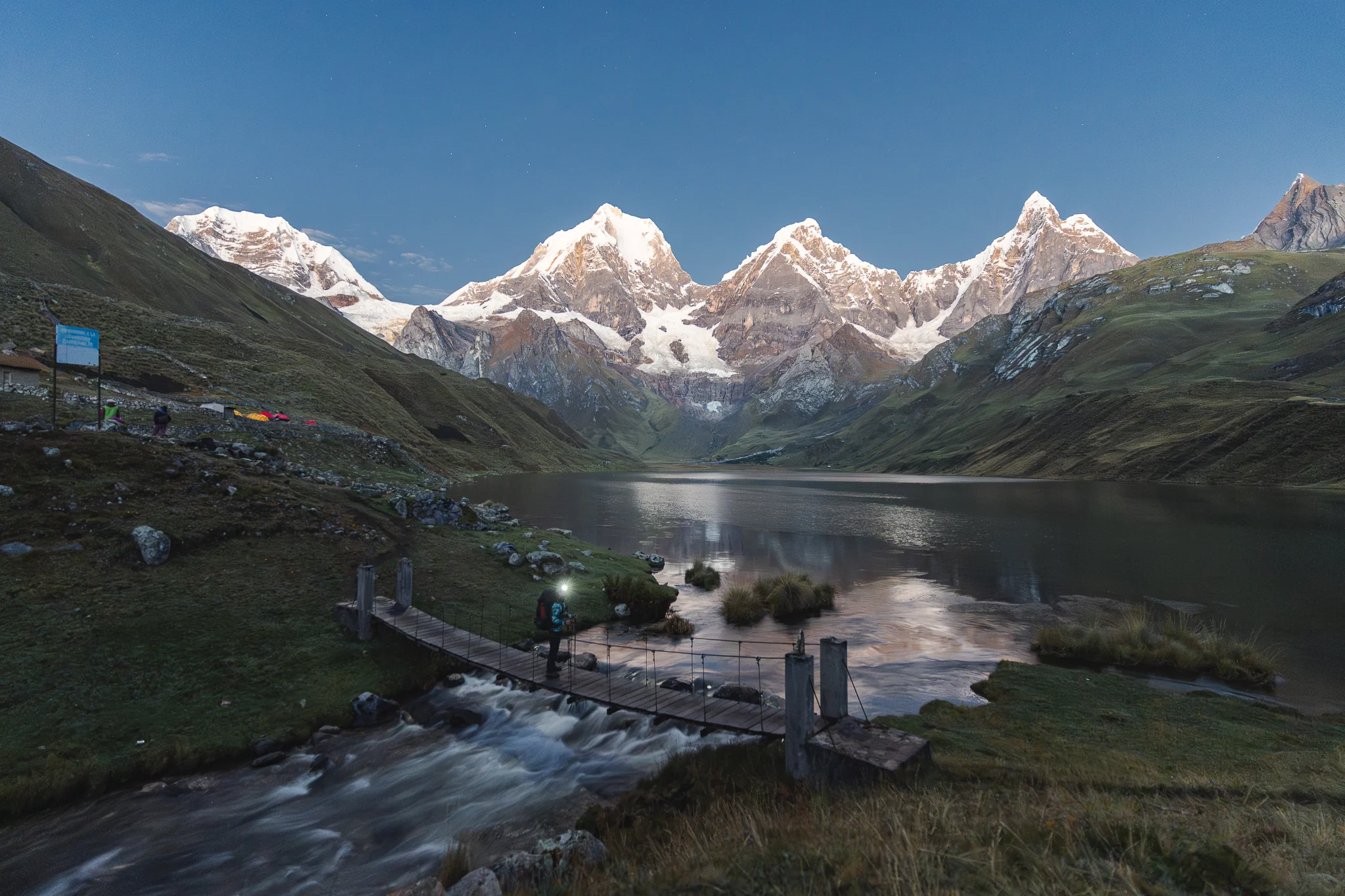 A person with a headlamp crossing a wooden footbridge over a stream at dawn, tents beside Laguna Carhuacocha and snow peaks lit pink beyond, Cordillera Huayhuash, Peru