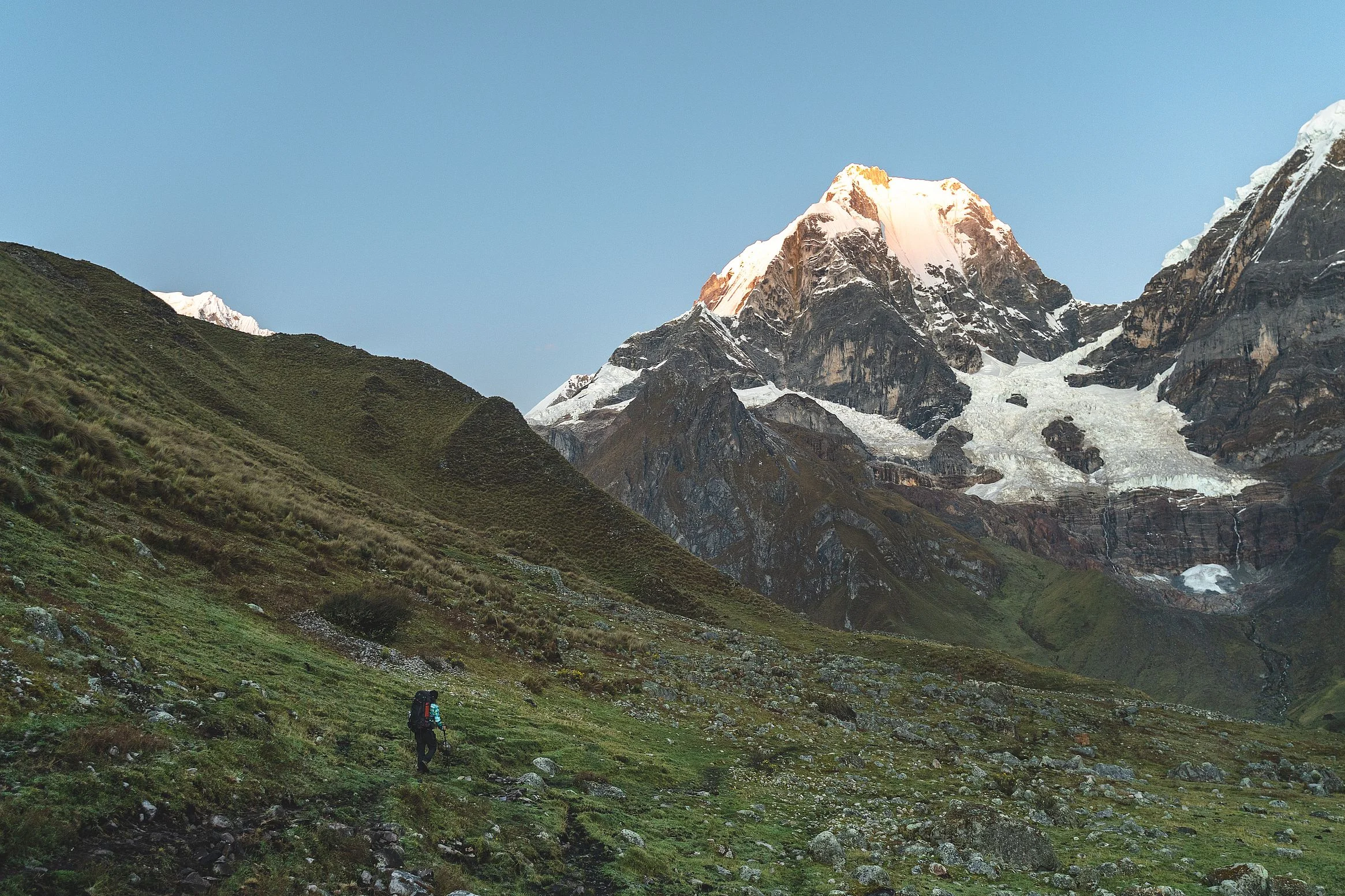 A trekker walking a green valley below the glaciers of Nevado Yerupajá, its summit lit orange by the last of the sun, Cordillera Huayhuash, Peru