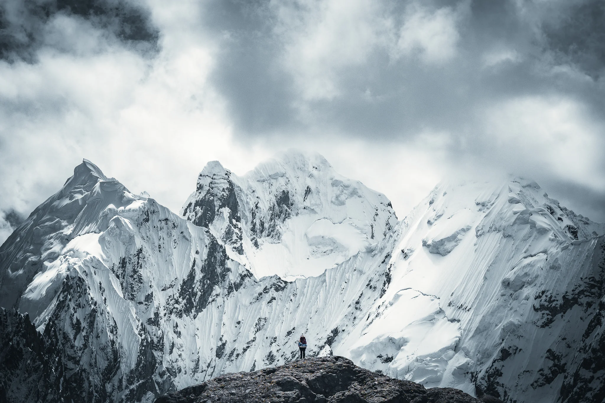 A small figure standing on a rocky outcrop in front of steep fluted snow and ice faces beneath heavy cloud, Cordillera Huayhuash, Peru