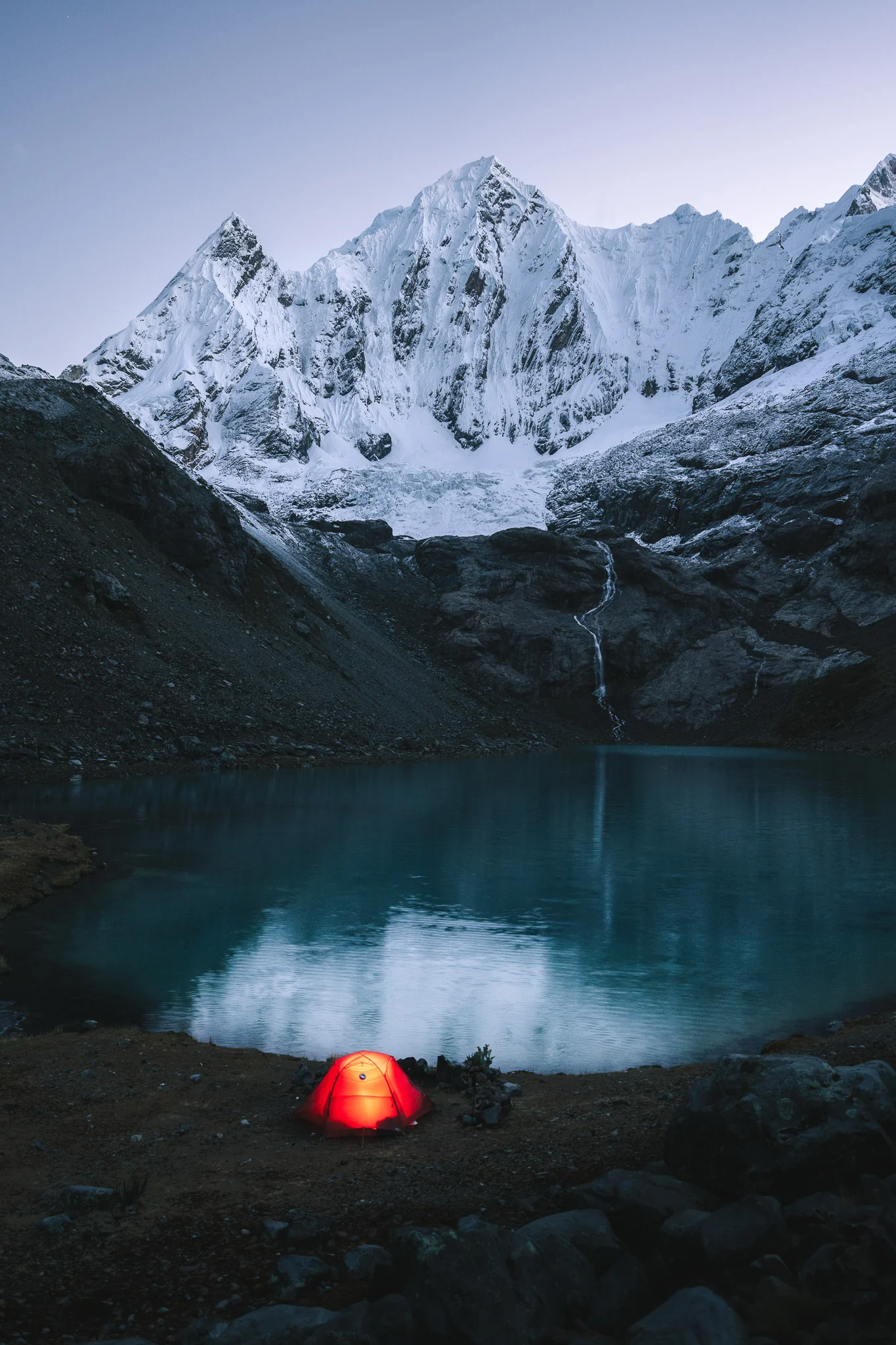 A glowing orange tent beside a dark glacial lake at twilight, a waterfall and snow-covered peaks rising behind, Cordillera Huayhuash, Peru