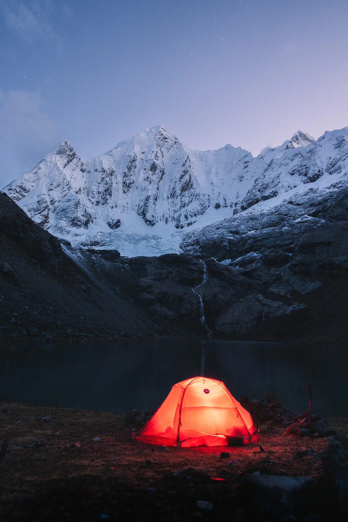 An orange tent glowing at night beside a lake, a waterfall and snow-covered peaks under a deep blue sky with faint stars, Cordillera Huayhuash, Peru