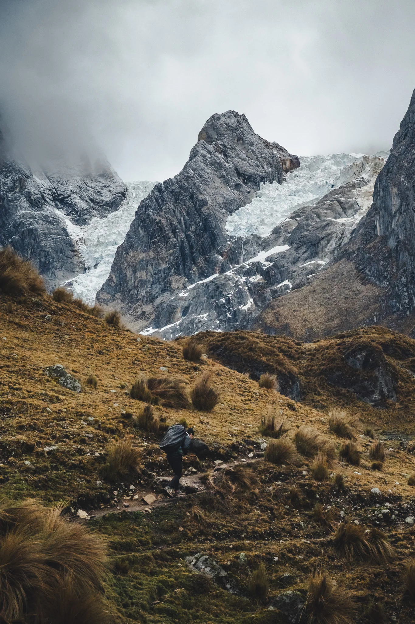 A trekker on a path through tussock grass below a glacier hanging between dark rock towers, cloud low over the peaks, Cordillera Huayhuash, Peru
