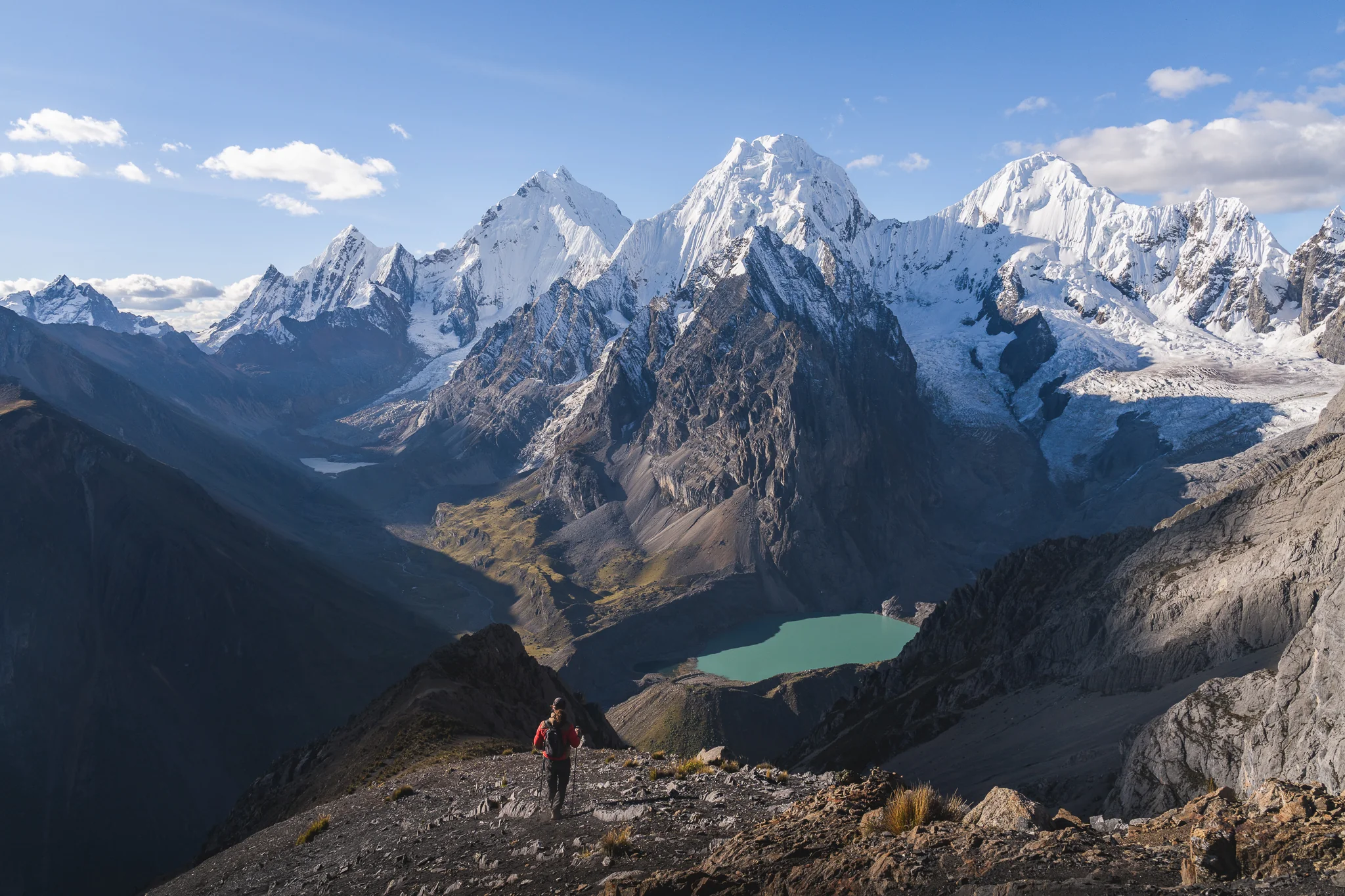 A hiker in a red jacket on a rocky high pass, a turquoise lake and a wall of glaciated peaks spread out behind, Cordillera Huayhuash, Peru