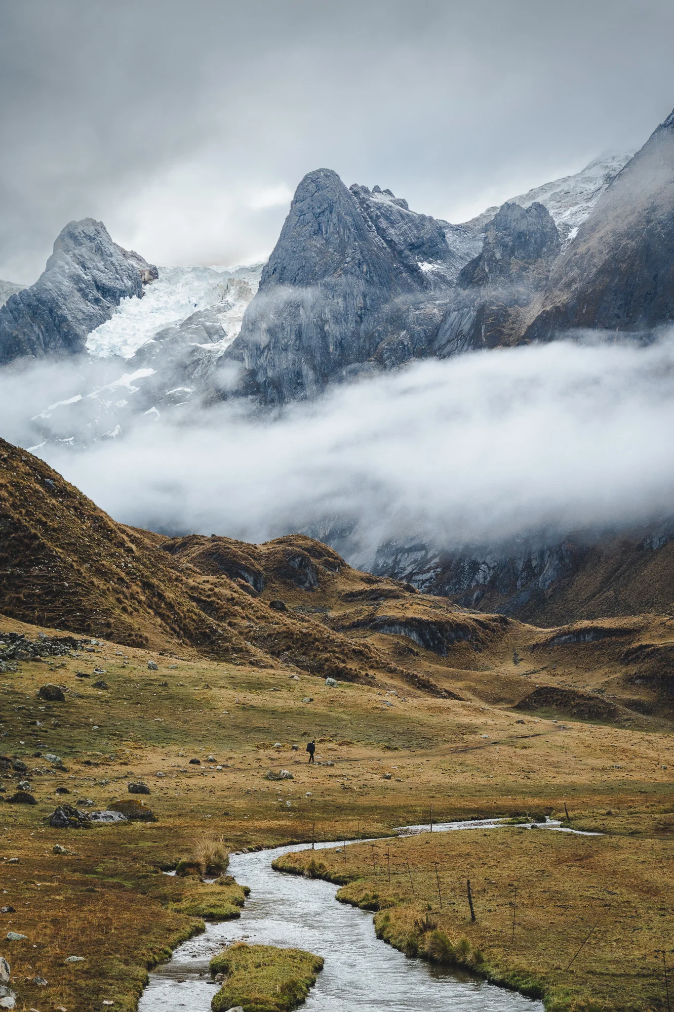 A trekker following a stream through a wide golden valley, cloud pouring off the glaciated peaks above, Cordillera Huayhuash, Peru