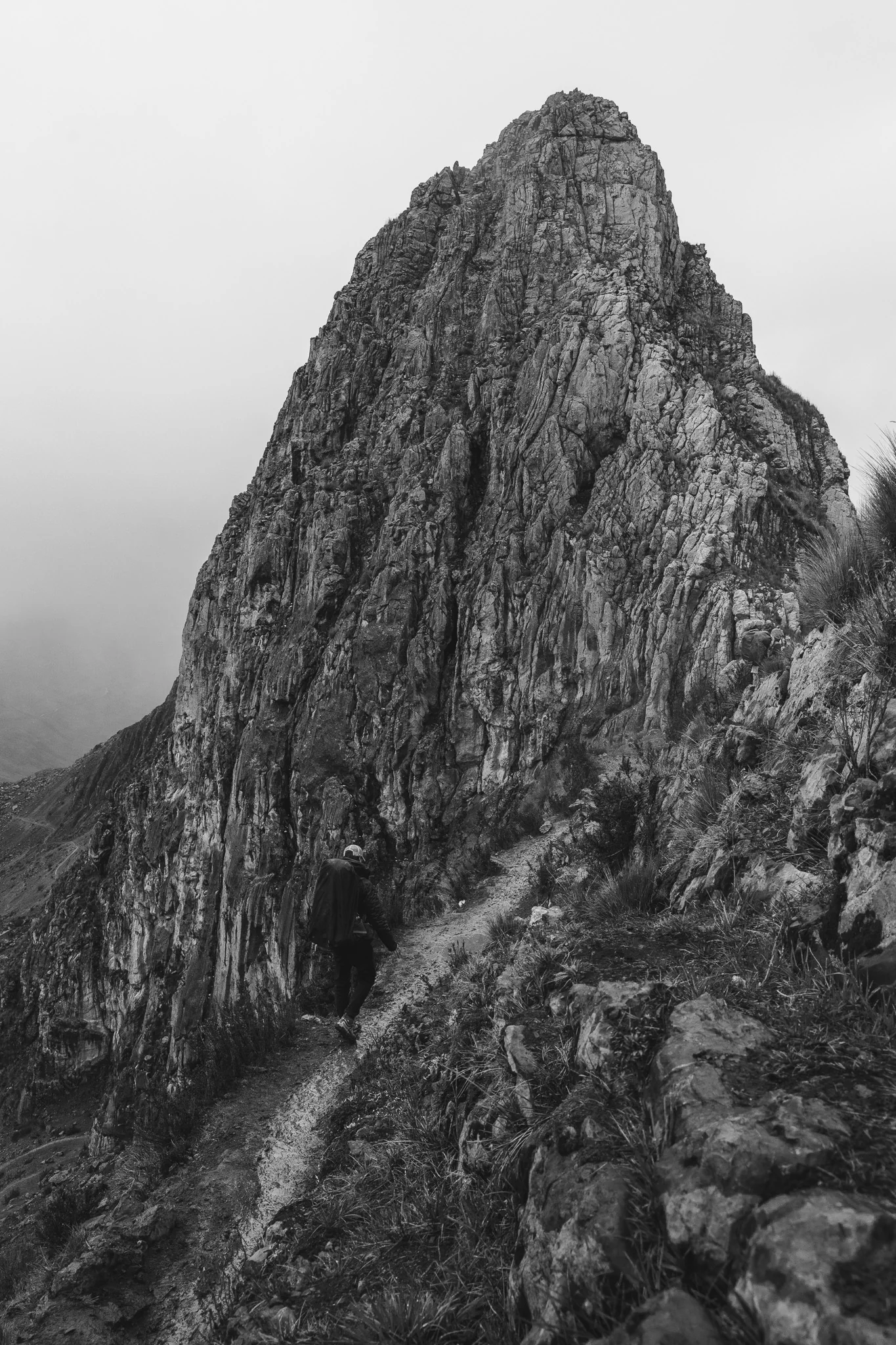 Black-and-white photo of a trekker on a narrow path beneath a huge fin of rock in the mist, first pass of the Huayhuash trek, Peru