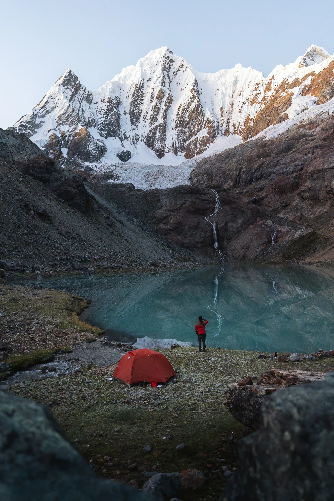 A red tent pitched on the grass beside the turquoise water of Laguna Caramarca, a trekker at the shore and the glaciers of Nevado Rasac behind, Peru