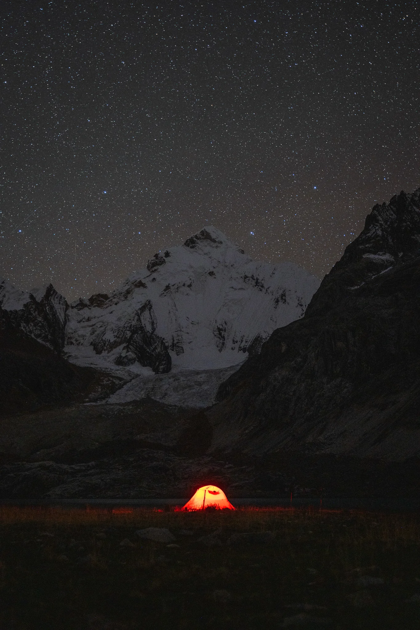 A tent glowing orange in the dark beside Laguna Sarapacocha under a sky thick with stars, the snow face of Yerupajá Sur rising behind, Peru