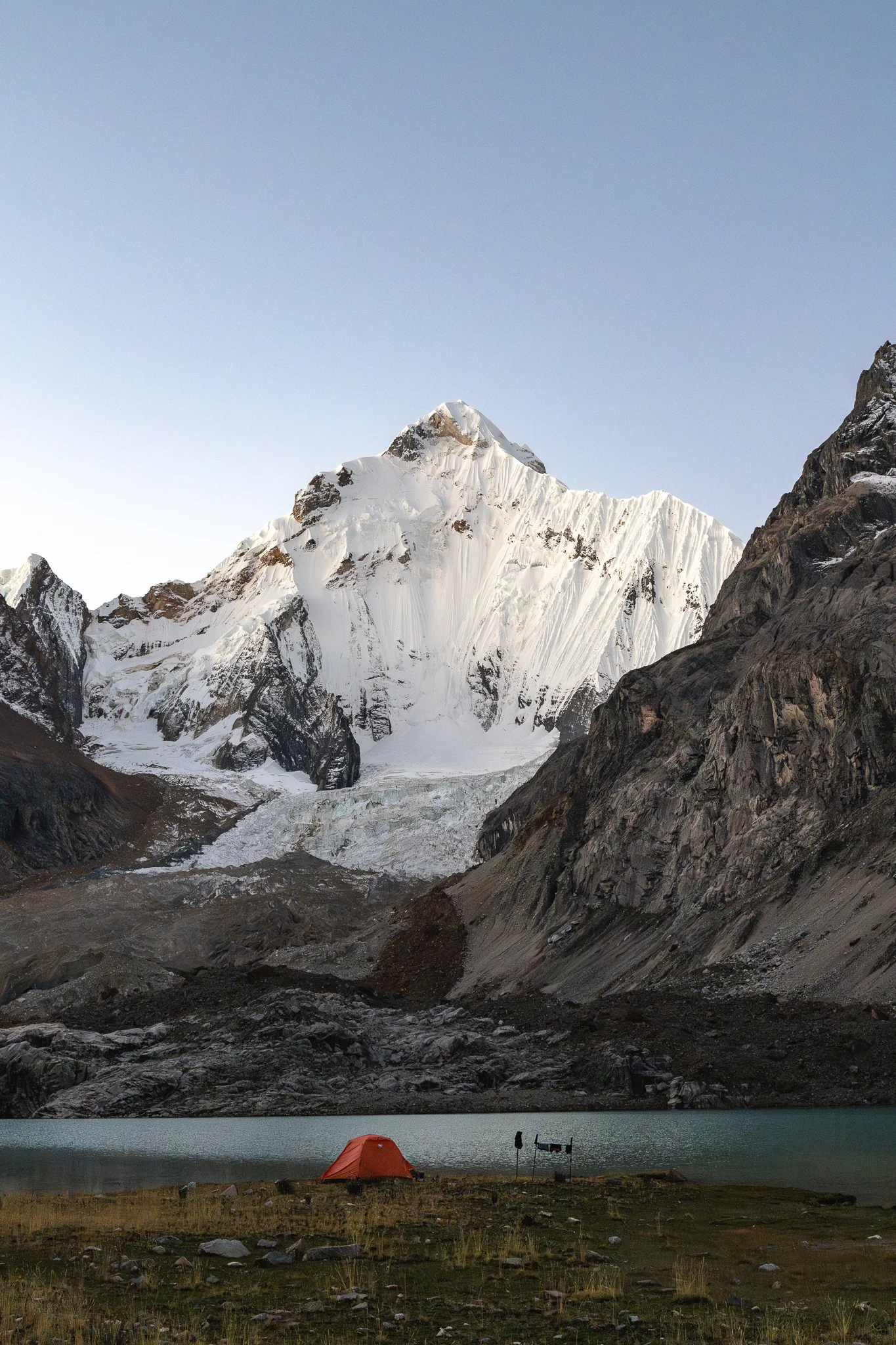 An orange tent on the shore of Laguna Sarapacocha in daylight, the glacier and snow face of Yerupajá Sur filling the head of the valley, Peru