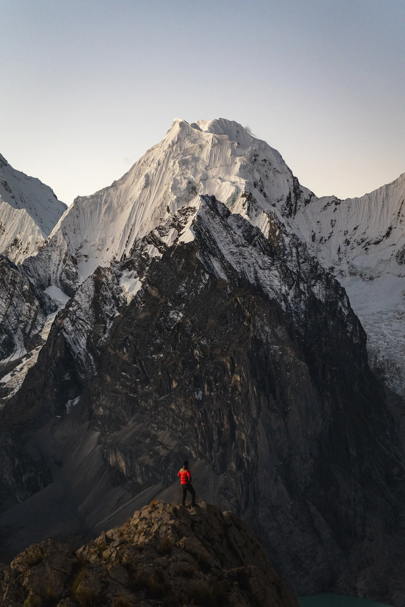 A trekker in a red jacket standing on a sharp ridge crest, a glaciated peak rising directly behind, near San Antonio pass, Cordillera Huayhuash, Peru