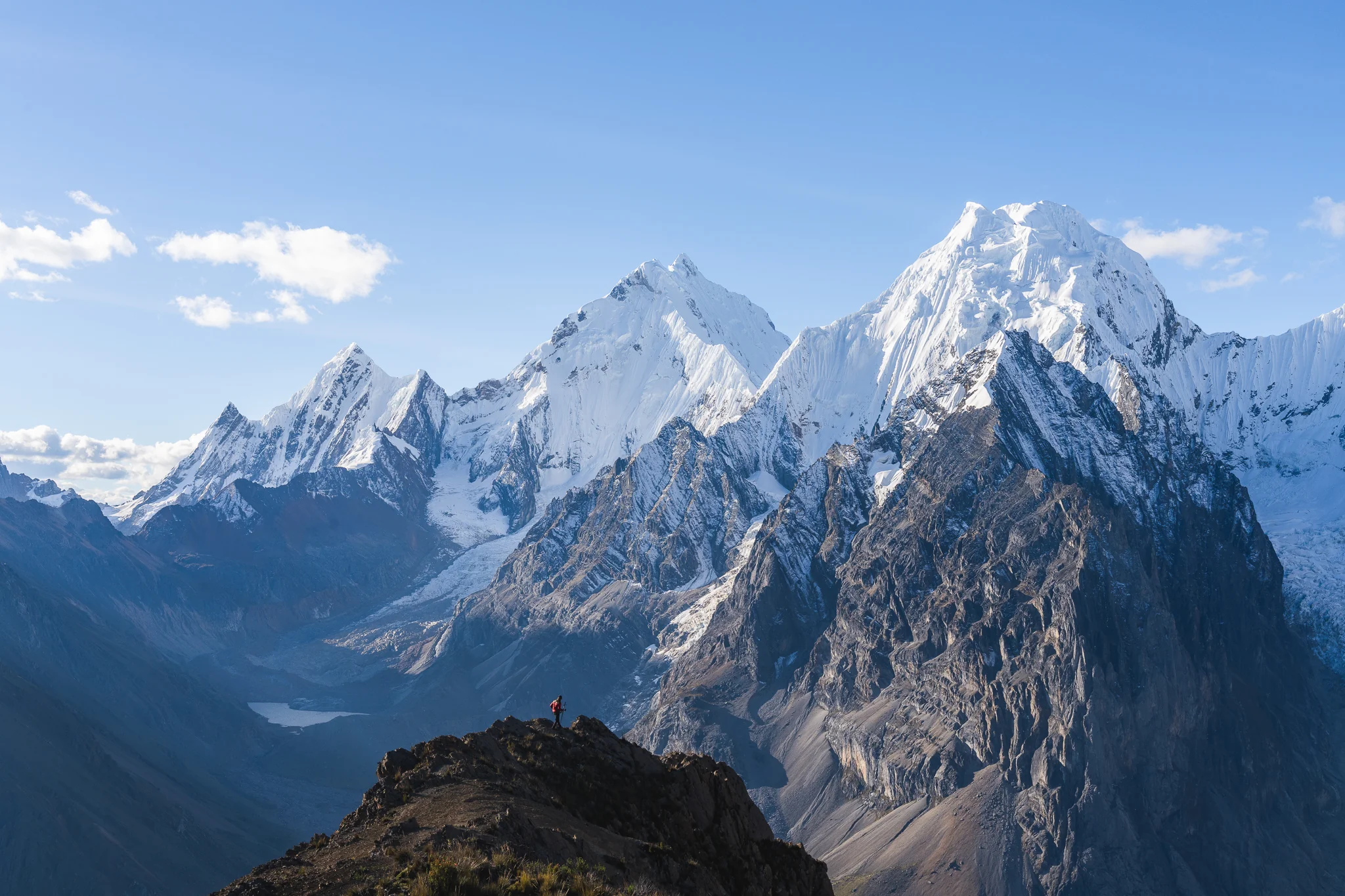A tiny figure on a rocky ridge above a lake, a wall of glaciated peaks lit by evening sun beyond, near San Antonio pass, Cordillera Huayhuash, Peru