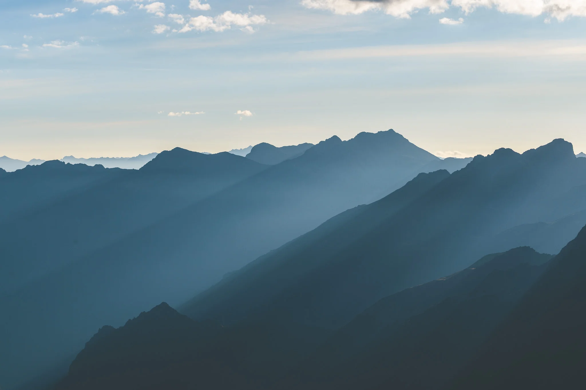 Shafts of sunlight through haze across layered blue mountain ridges under a pale sky, Cordillera Huayhuash, Peru